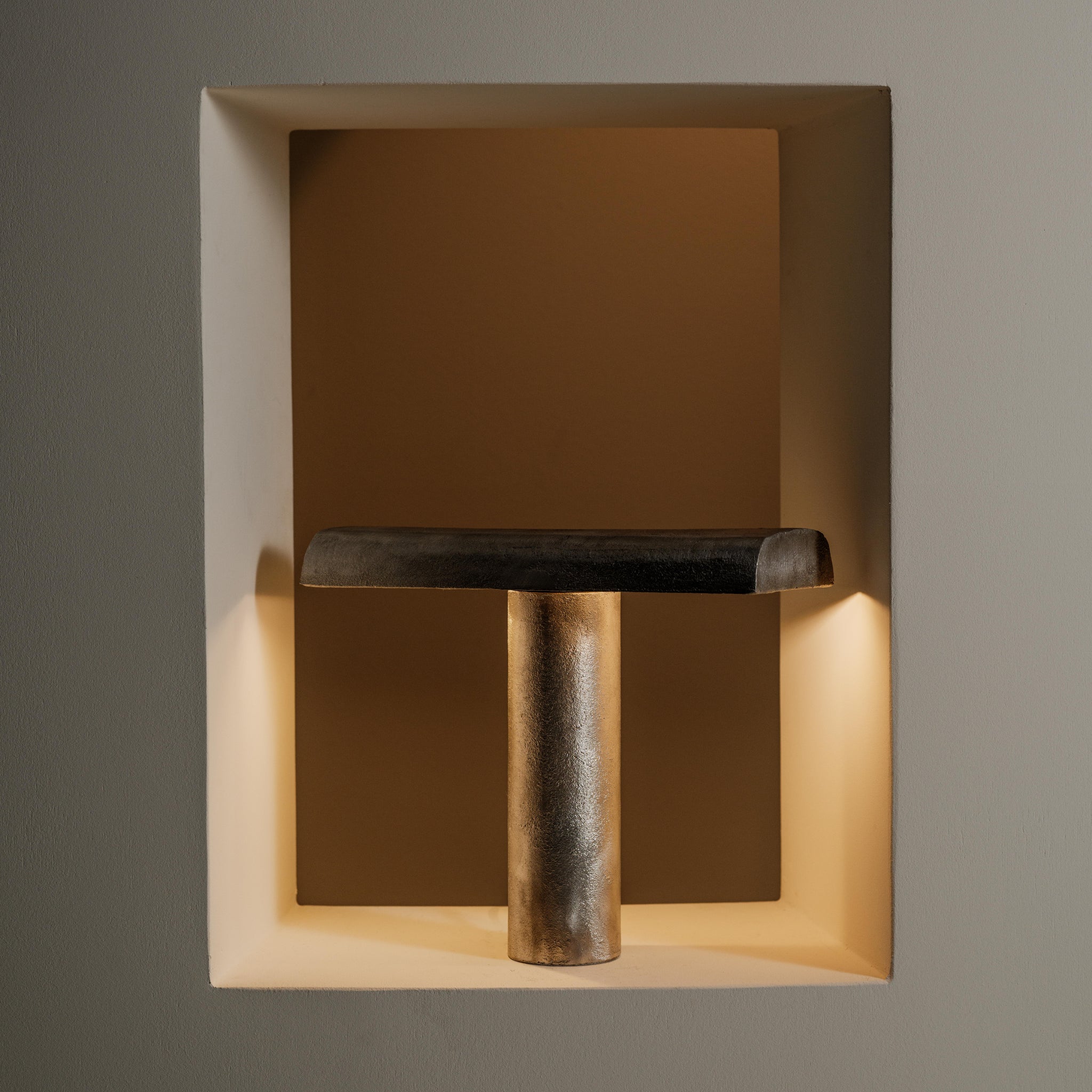 De Serax Fck Tafellamp Staal Grijs van Serax, een opvallende metalen T-vormige lamp, wordt tentoongesteld in een verzonken, zacht verlichte nis die de gladde textuur en het minimalistische ontwerp, geïnspireerd door brutalistische architectuur, benadrukt.