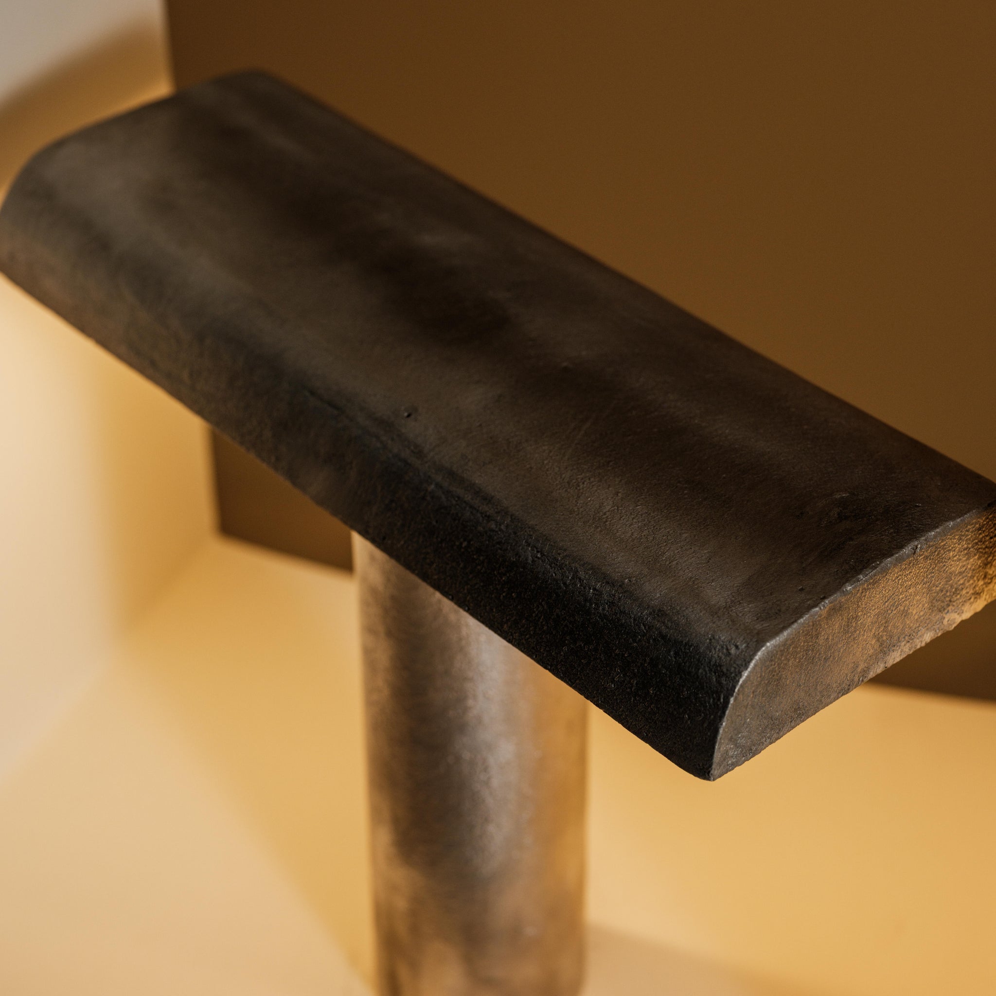 Een close-up van de Serax Fck Tafellamp Staal Grijs van Serax, een T-vormige staalgrijze tafellamp met brutalistisch ontwerp, tegen een beige en bruine achtergrond met zachte verlichting.