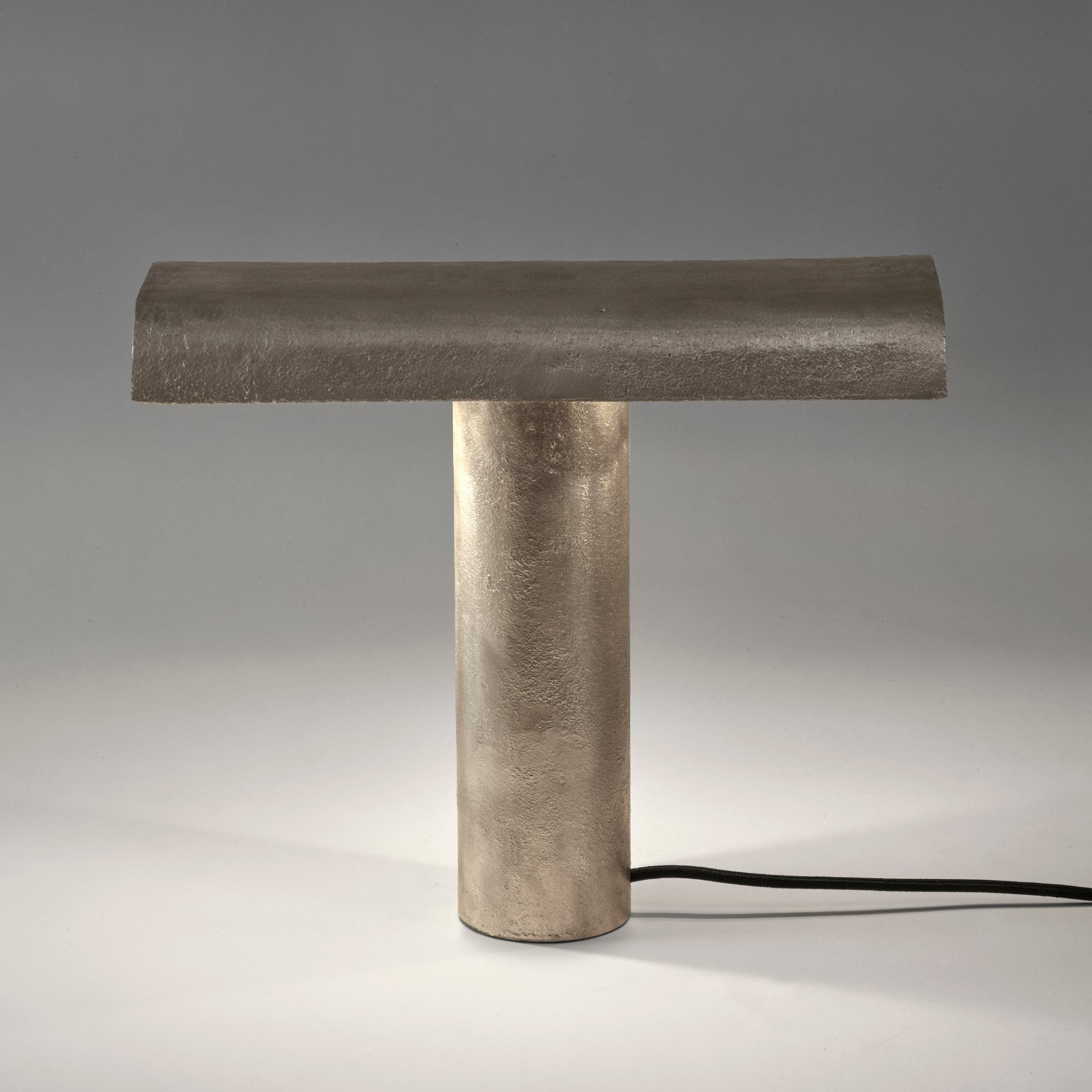 De Serax Fck Tafellamp Staal Grijs van Serax heeft een modern, minimalistisch ontwerp met een cilindrische voet, platte rechthoekige bovenkant, afwerking met metallic textuur en zichtbaar zwart netsnoer op een wit oppervlak.