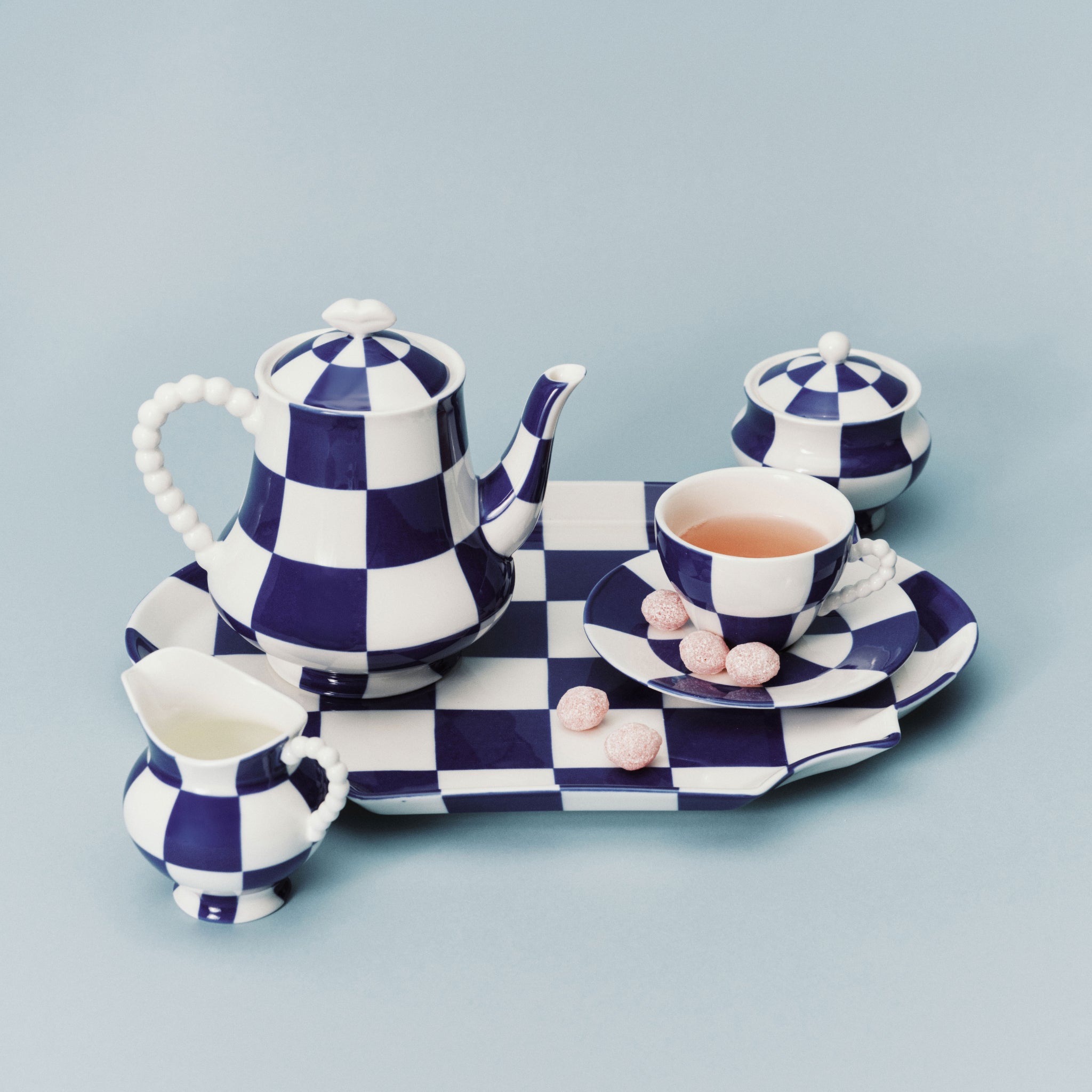 Het Serax Wouters & Hendrix Fête Des Bêtes Melkkannetje Blauw Wit wordt getoond als onderdeel van een blauw-wit geblokt Serax theeservies, gerangschikt met roze snoepjes op het schoteltje tegen een lichtblauwe achtergrond.