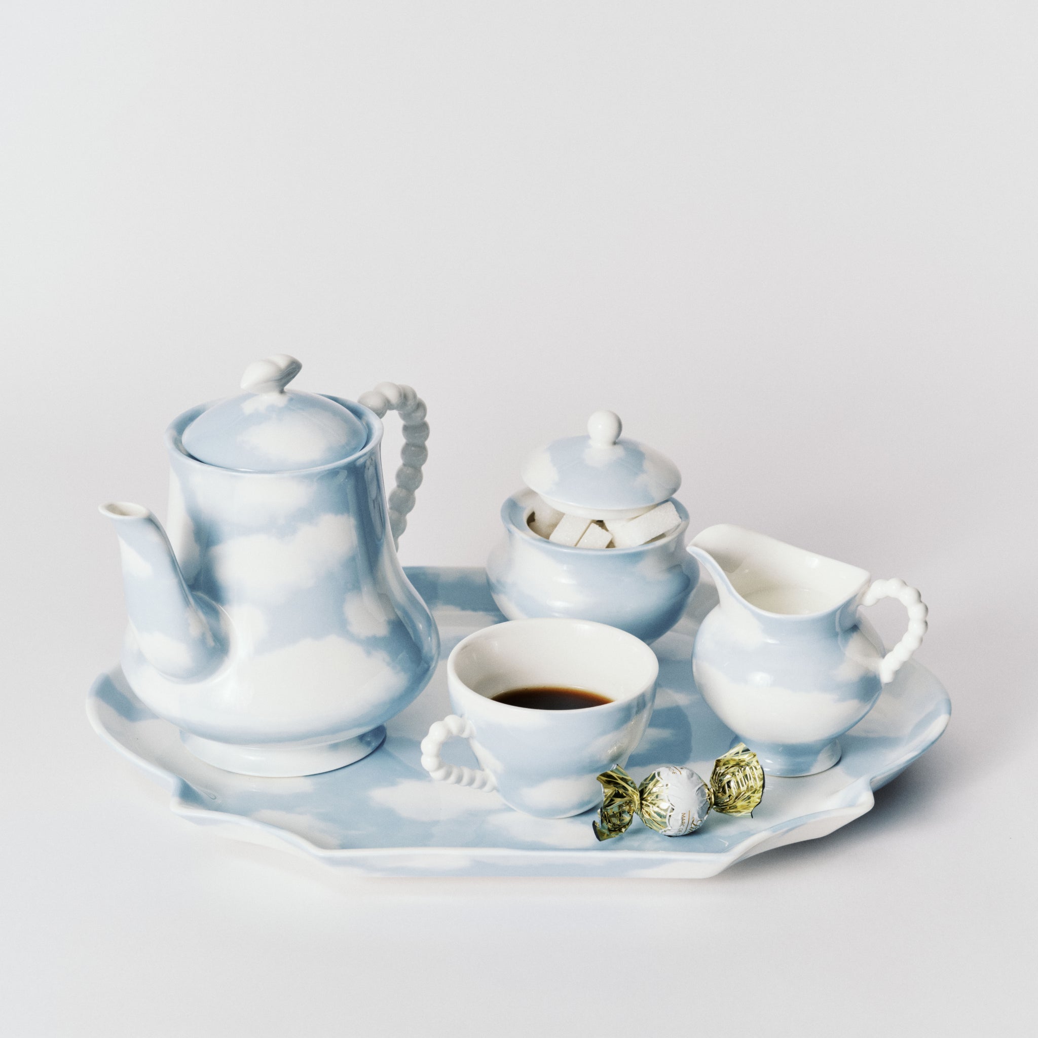 De Serax Wouters & Hendrix Fête Des Bêtes Koffiekop + Schotel Lichtblauw heeft een blauw-wit wolkenluchtontwerp, uitgestald met koffie en verpakte snoepjes op een bijpassend dienblad tegen een witte achtergrond.