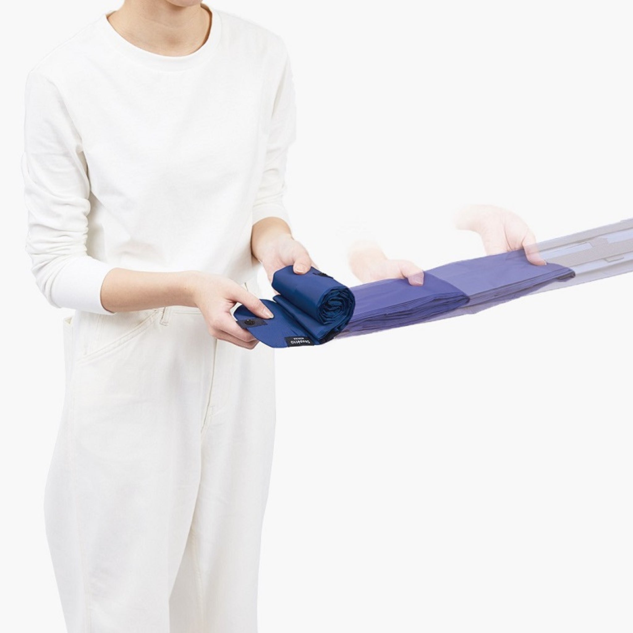 Een persoon in een witte outfit vouwt een donkergroene Shupatto Opvouwbare Boodschappentas L van Shupatto, met bewegingsonscherpte die laat zien hoe de stof stap voor stap wordt gevouwen.