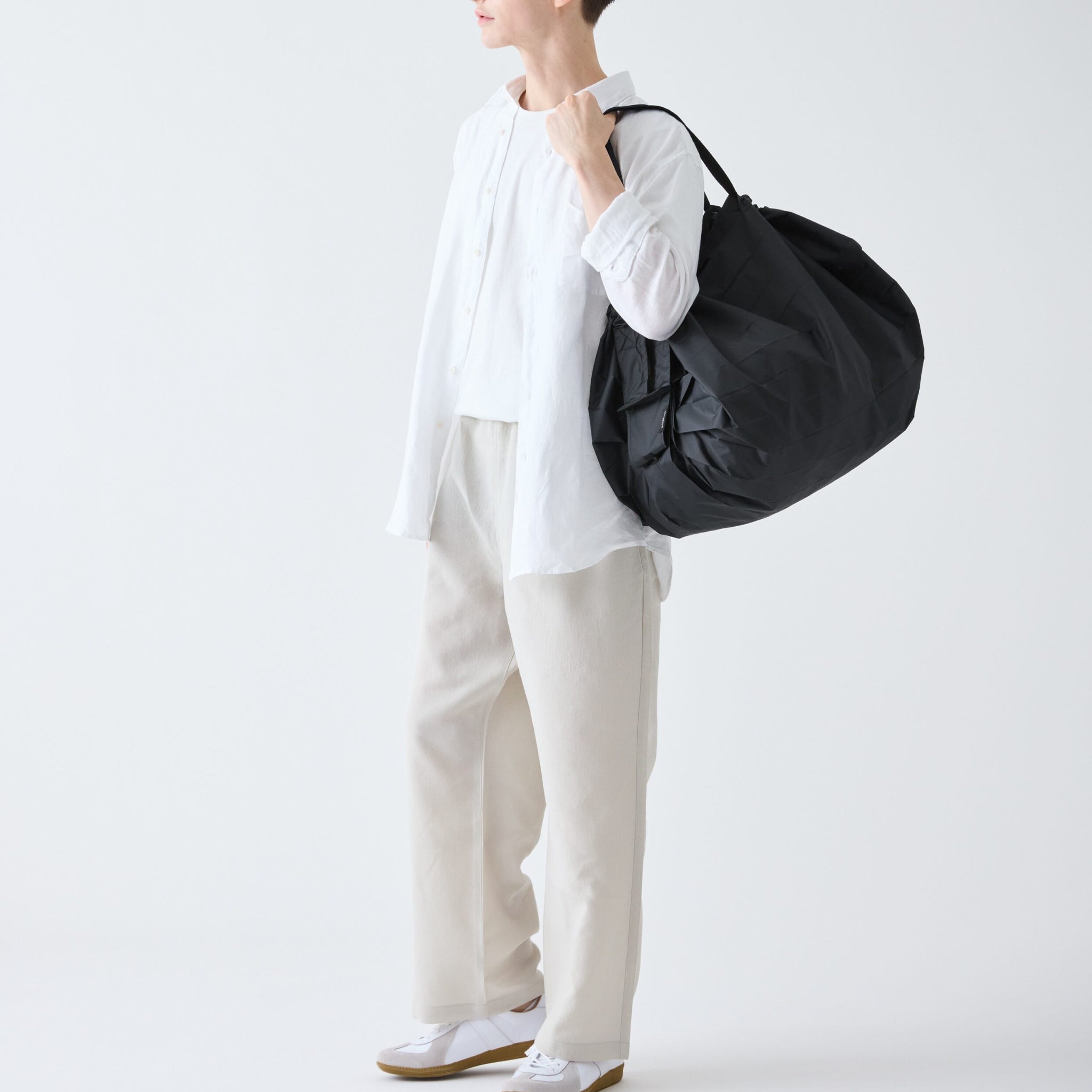 Een persoon in een wit shirt, lichte broek en witte sneakers staat tegen een effen achtergrond met een Shupatto Opvouwbare Boodschappentas Jet Zwart L van Shupatto over zijn schouder. Hun gezicht is gedeeltelijk buiten beeld.