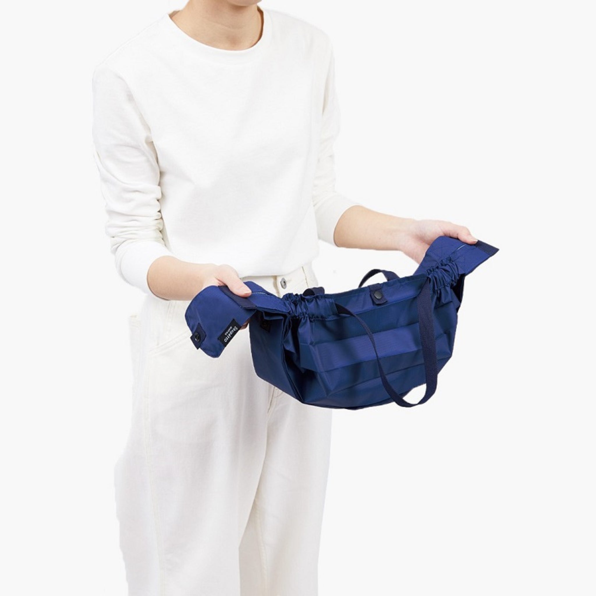 Een persoon in een wit shirt met lange mouwen en een broek houdt een open Shupatto Opvouwbare Boodschappentas Mori Groen M tegen een effen witte achtergrond.