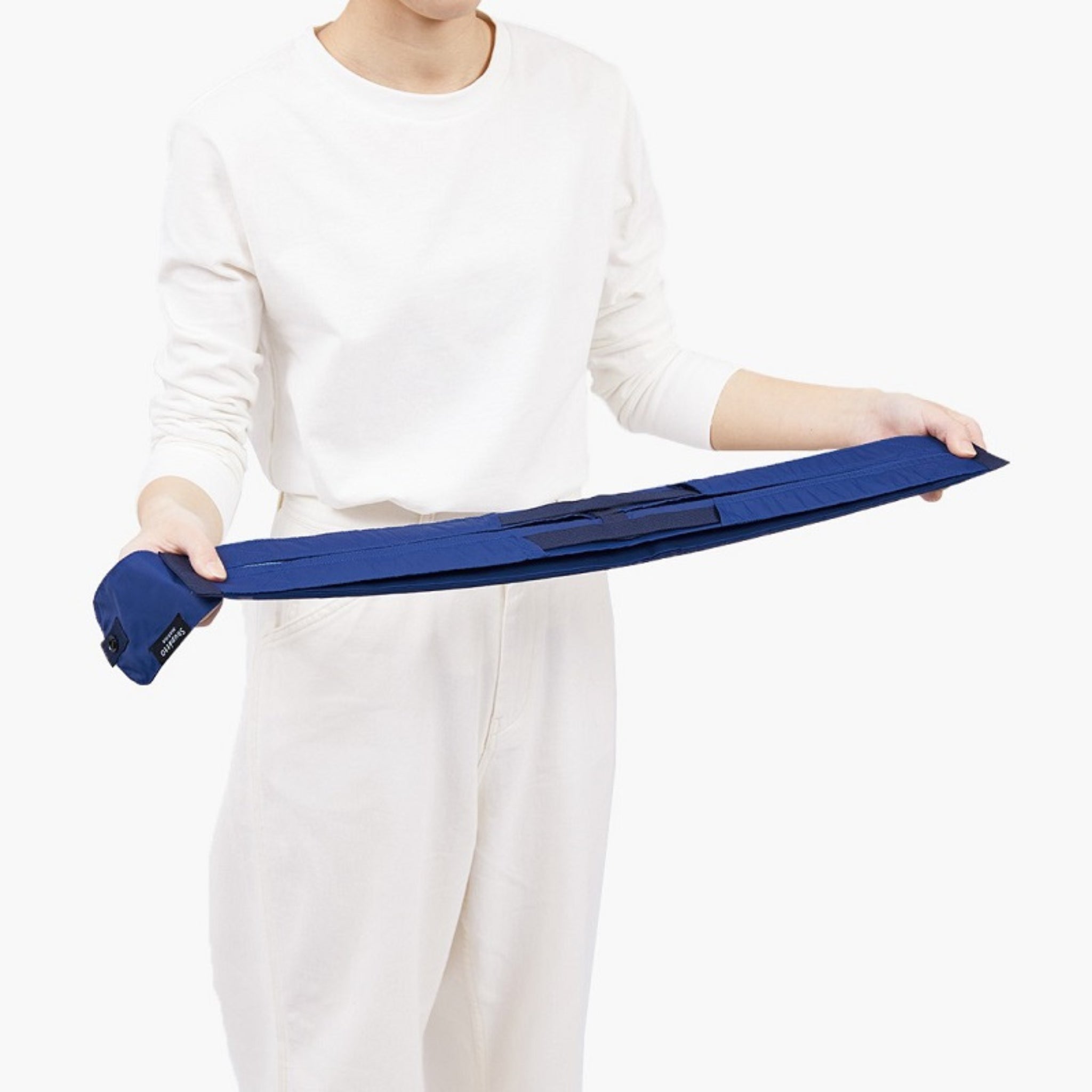 Een persoon in een witte outfit houdt een Shupatto Opvouwbare Boodschappentas Mori Groen M in beide handen tegen een effen witte achtergrond. Dit toont het compacte, gestructureerde ontwerp van deze rechthoekige Shupatto tas.