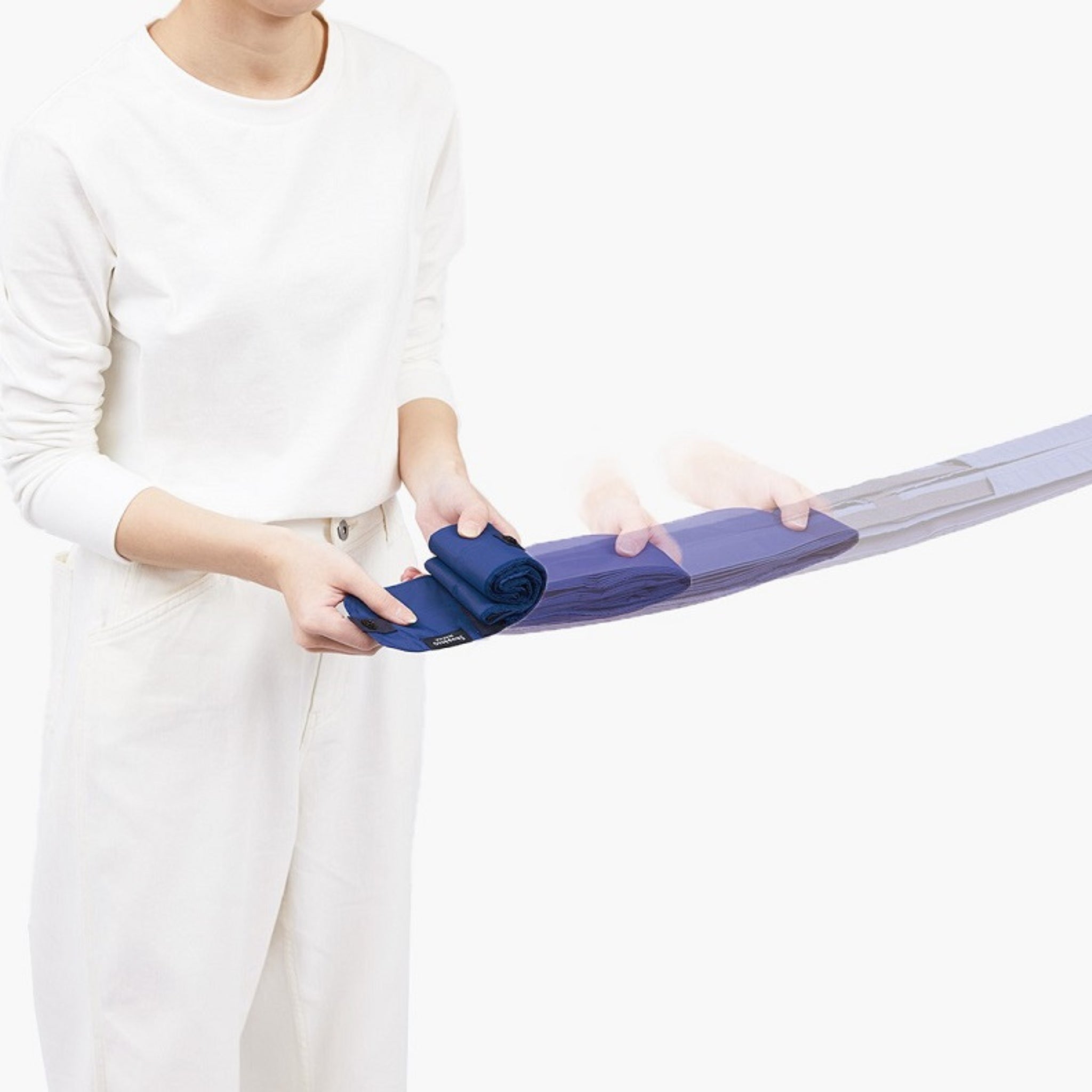 Een persoon in een volledig witte outfit demonstreert het opvouwen van de Shupatto Opvouwbare Boodschappentas Cashew Nuts Beige M, met bewegingsonscherpte die het gemakkelijk opvouwen benadrukt. Een effen witte achtergrond houdt de aandacht gericht op het vouwproces van de Shupatto tas.