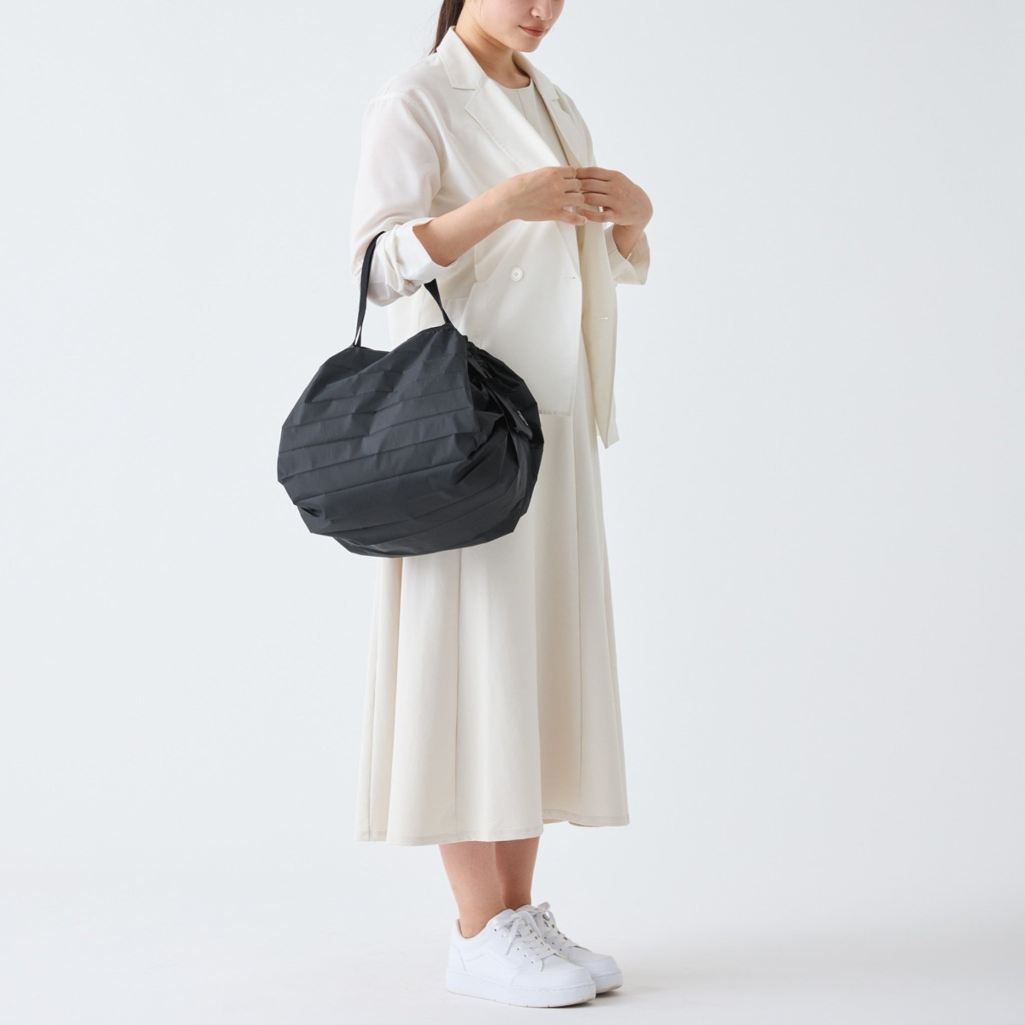 Een vrouw in een witte jurk en sneakers staat tegen een effen achtergrond en houdt de Shupatto Opvouwbare Boodschappentas Jet Zwart M van Shupatto vast. Haar hoofd helt naar beneden terwijl haar handen rusten naast de riem van de herbruikbare tas.