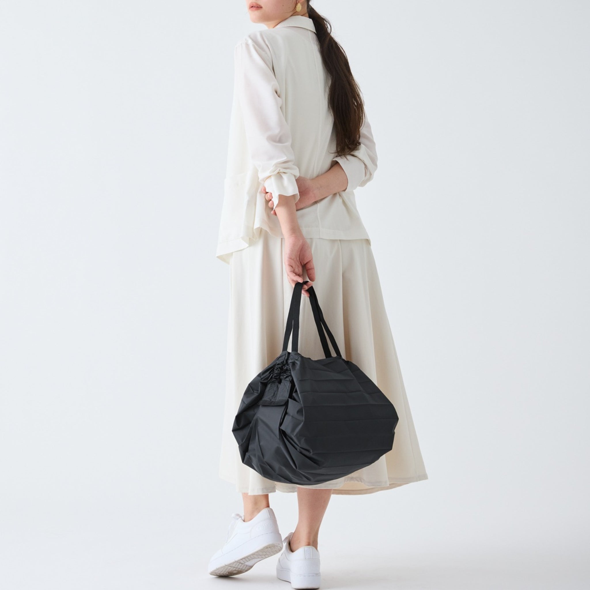 Een vrouw in een off-white jurk met lange mouwen en witte sneakers staat met haar rug naar haar toe gekeerd en houdt een Shupatto Opvouwbare Boodschappentas Jet Zwart M van Shupatto vast tegen een effen witte achtergrond.