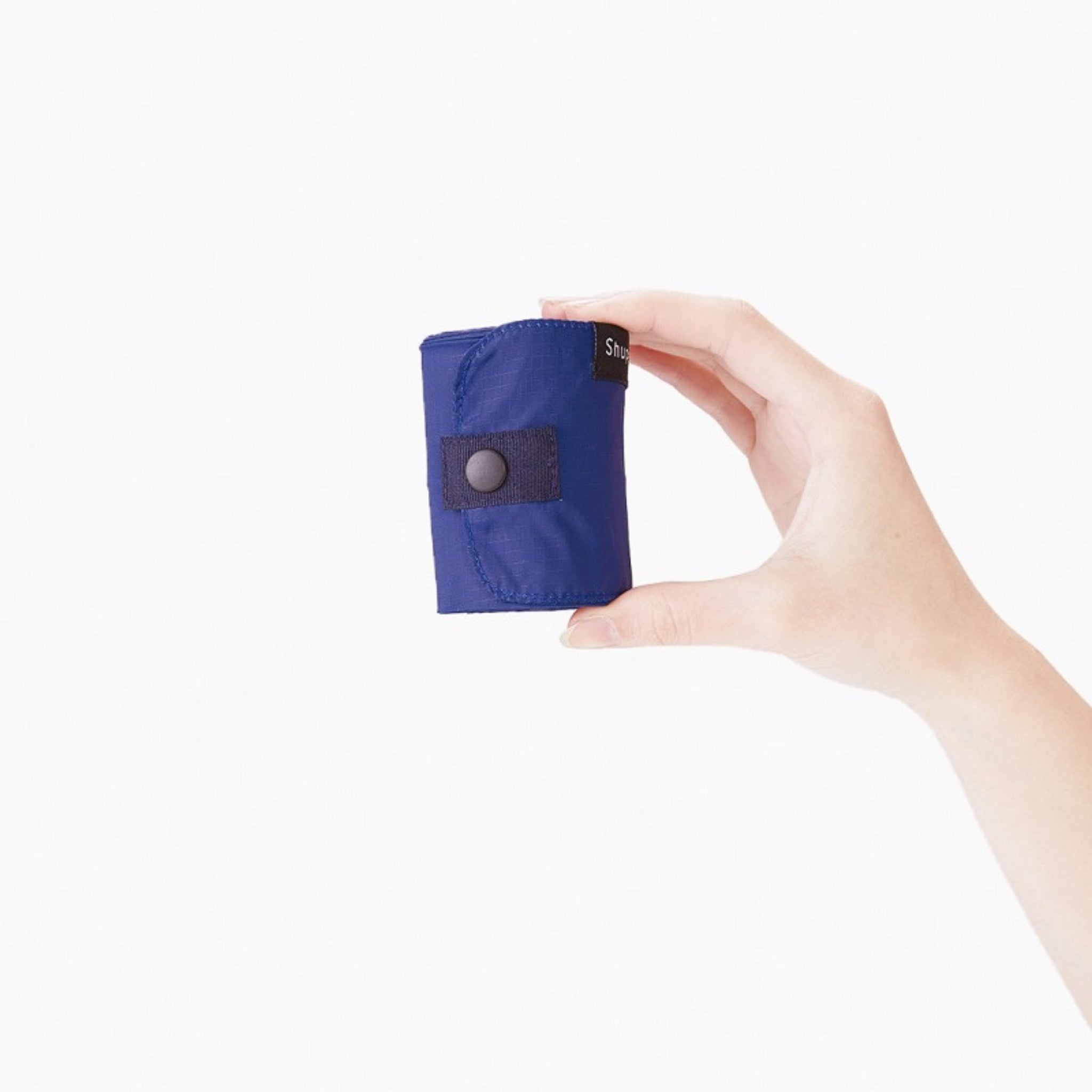Een hand houdt een compacte, opgerolde Shupatto Opvouwbare Boodschappentas Yoru Donkerblauw M met drukknoopsluiting van Shupatto tegen een effen witte achtergrond.