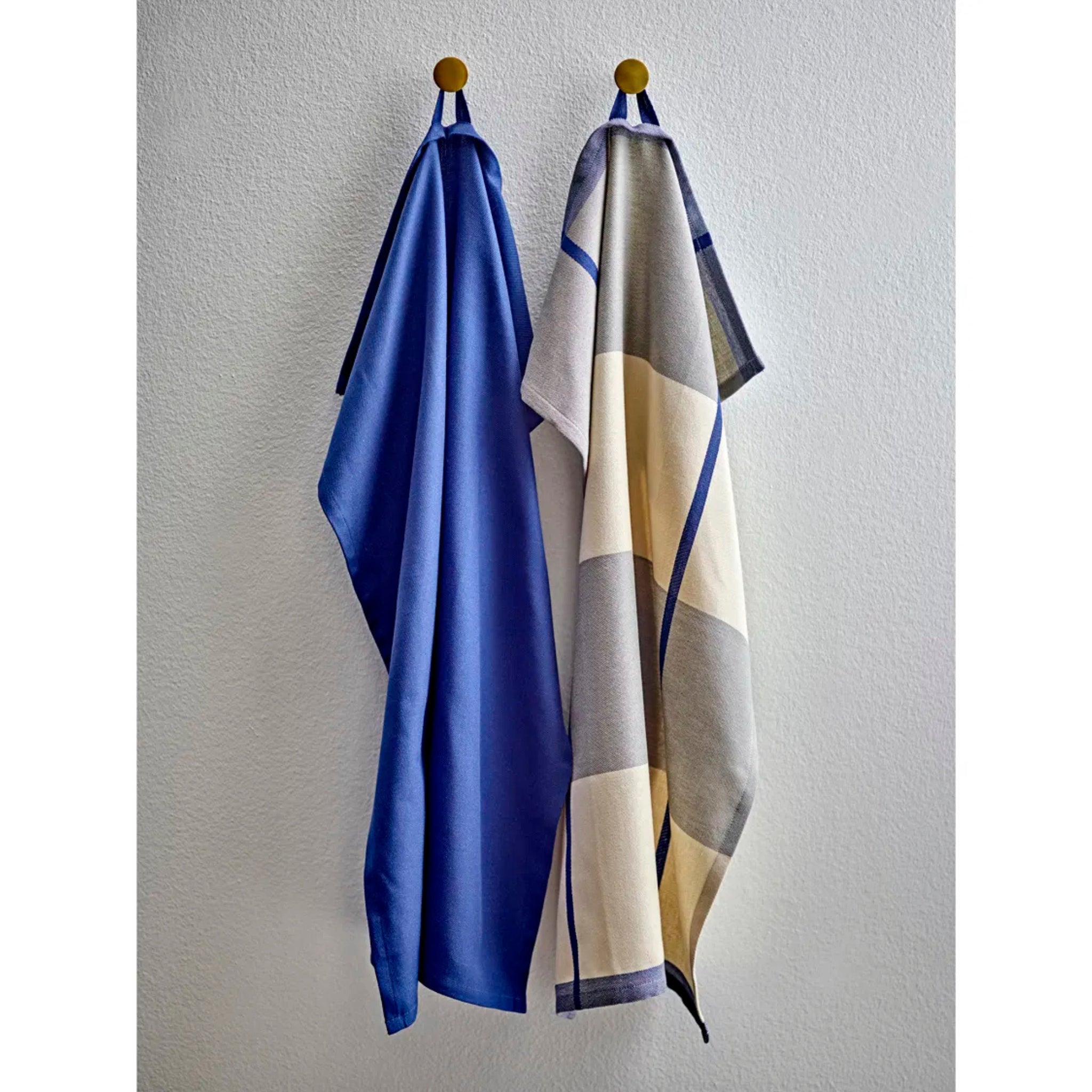 Twee handdoeken hangen aan ronde haken tegen een witte muur; de linker is effen blauw en de rechter is Södahl Blocks Theedoek Blauw-een stijlvolle keuze om Scandinavische flair aan je keuken toe te voegen. Set bestaat uit 2 handdoeken.