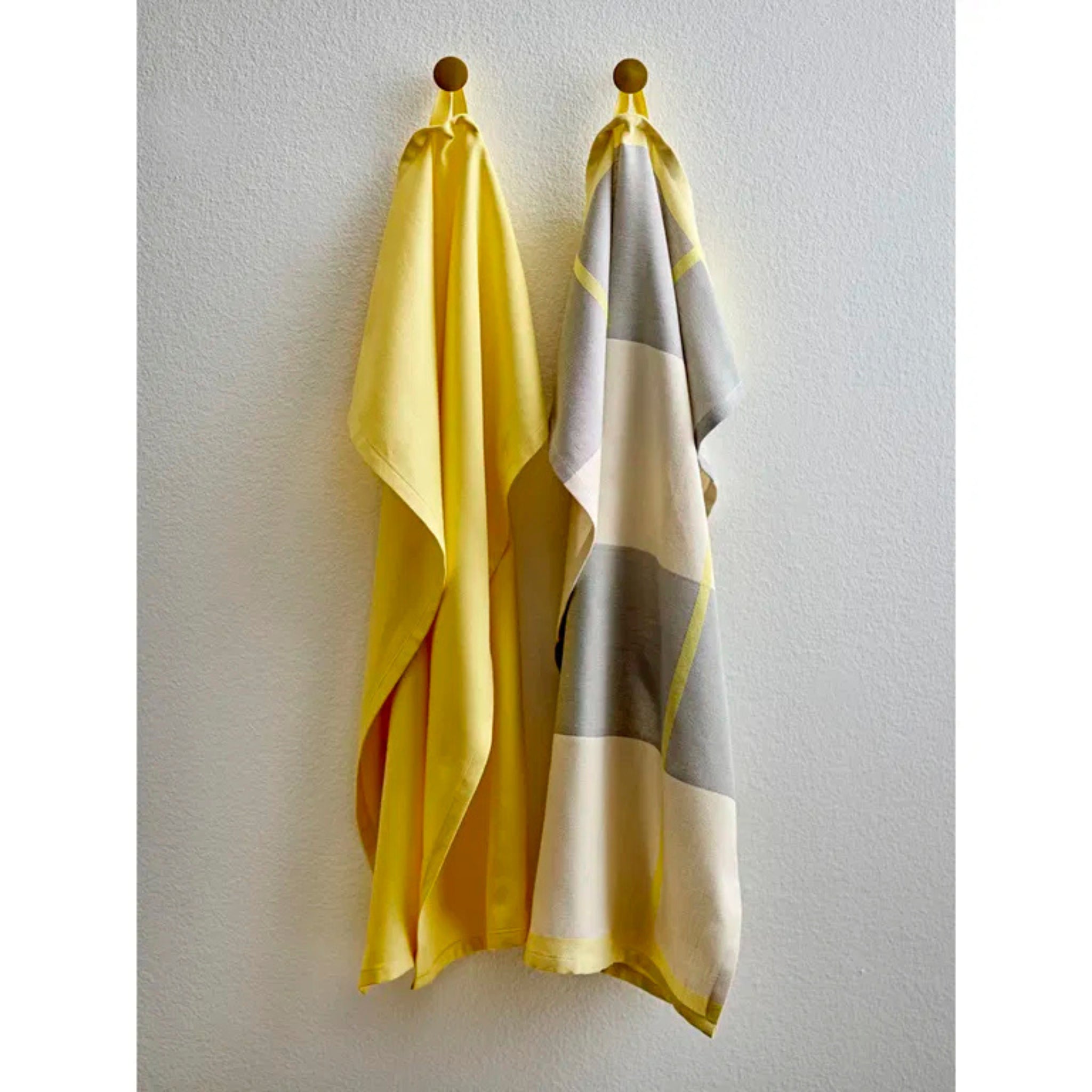 Twee Södahl Blocks Theedoeken Geel hangen aan wandhaken - de ene effen geel, de andere met een wit, grijs, beige en geel blokpatroon. Beide zijn van Södahl en hangen tegen een effen witte muur.