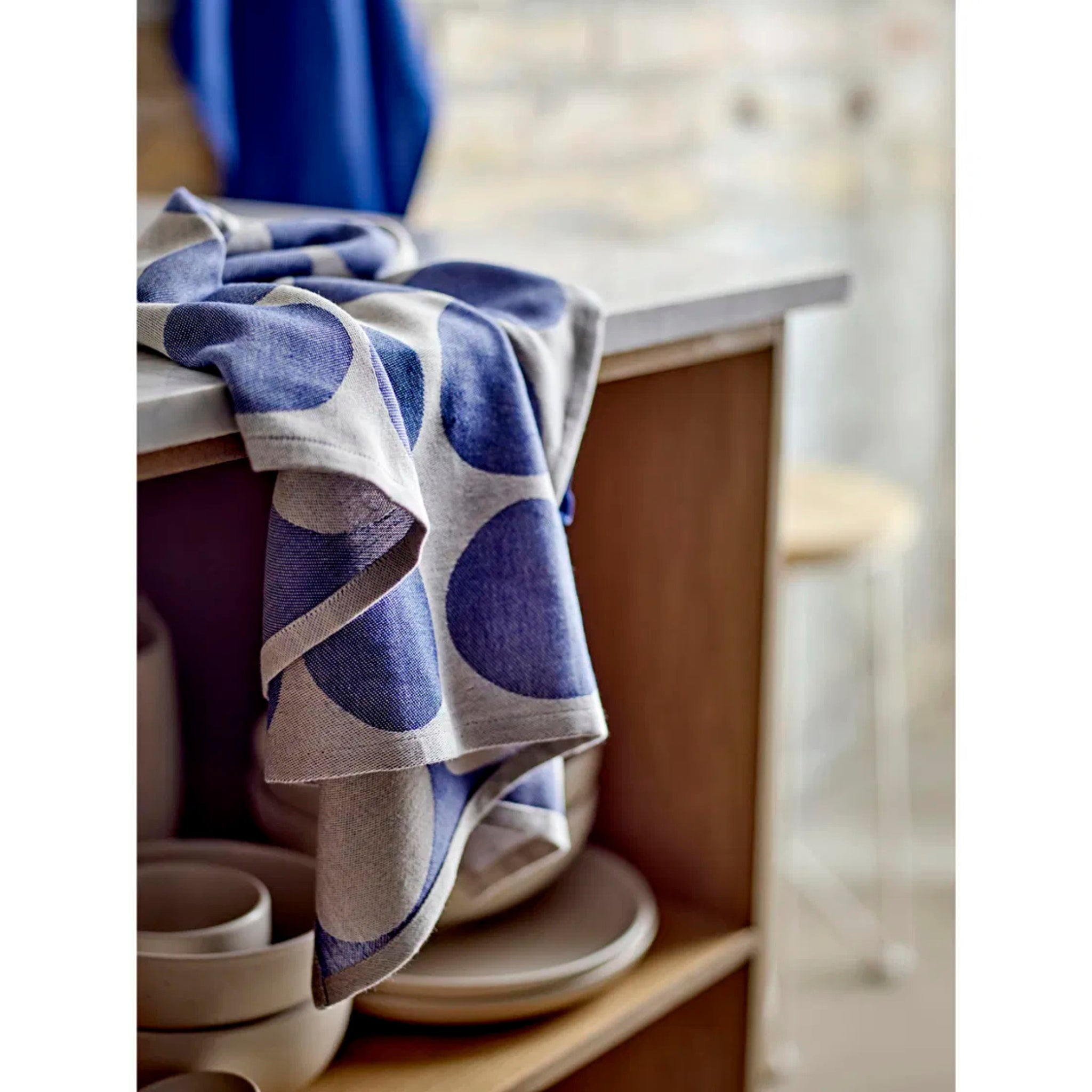 Een set van twee Södahl Bubbles Theedoeken in blauw, met een gedurfd stippenpatroon, hangt boven een houten keukenplank, met daaronder gestapelde kommen en borden van keramiek, wat het elegante Scandinavische design benadrukt.