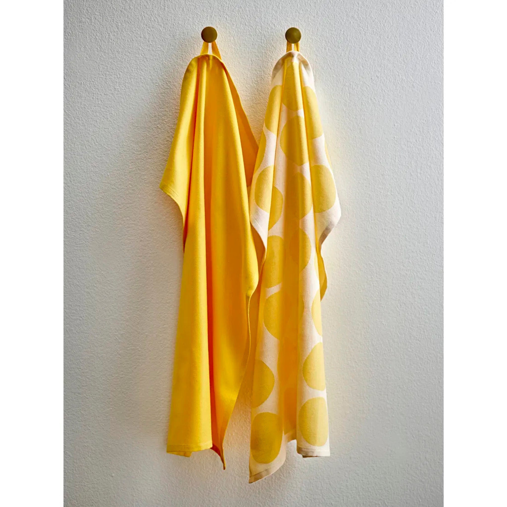 Twee gele handdoeken-Södahl Bubbles Theedoek Geel 2 st. - hangen aan wandhaken; de ene is effen, de andere toont grote gele cirkels op wit en brengt een Scandinavisch tintje in je keuken.