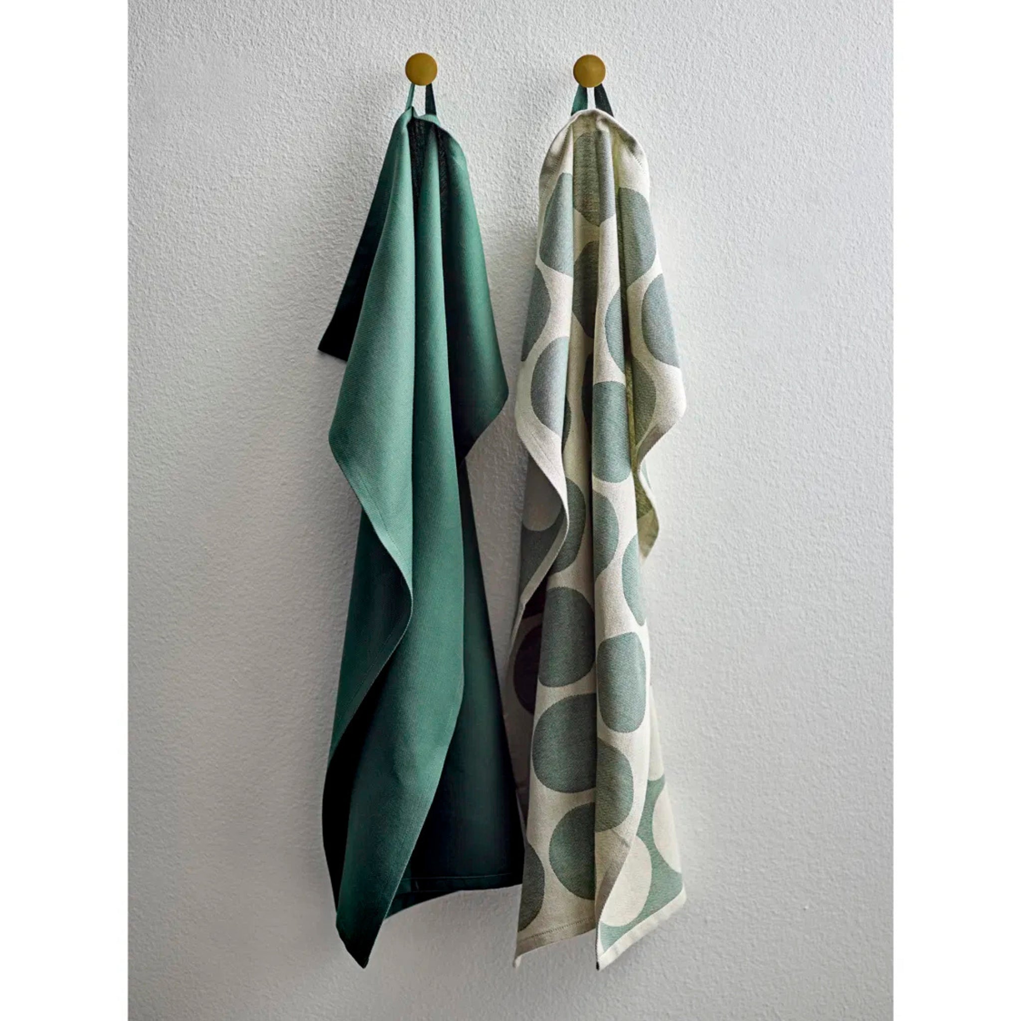 Twee handdoeken hangen aan ronde haken tegen een witte muur met structuur: links een groene theedoek, rechts een Södahl Bubbles Theedoek Groen 2 st. - een perfecte Scandinavische keukenaccessoire.