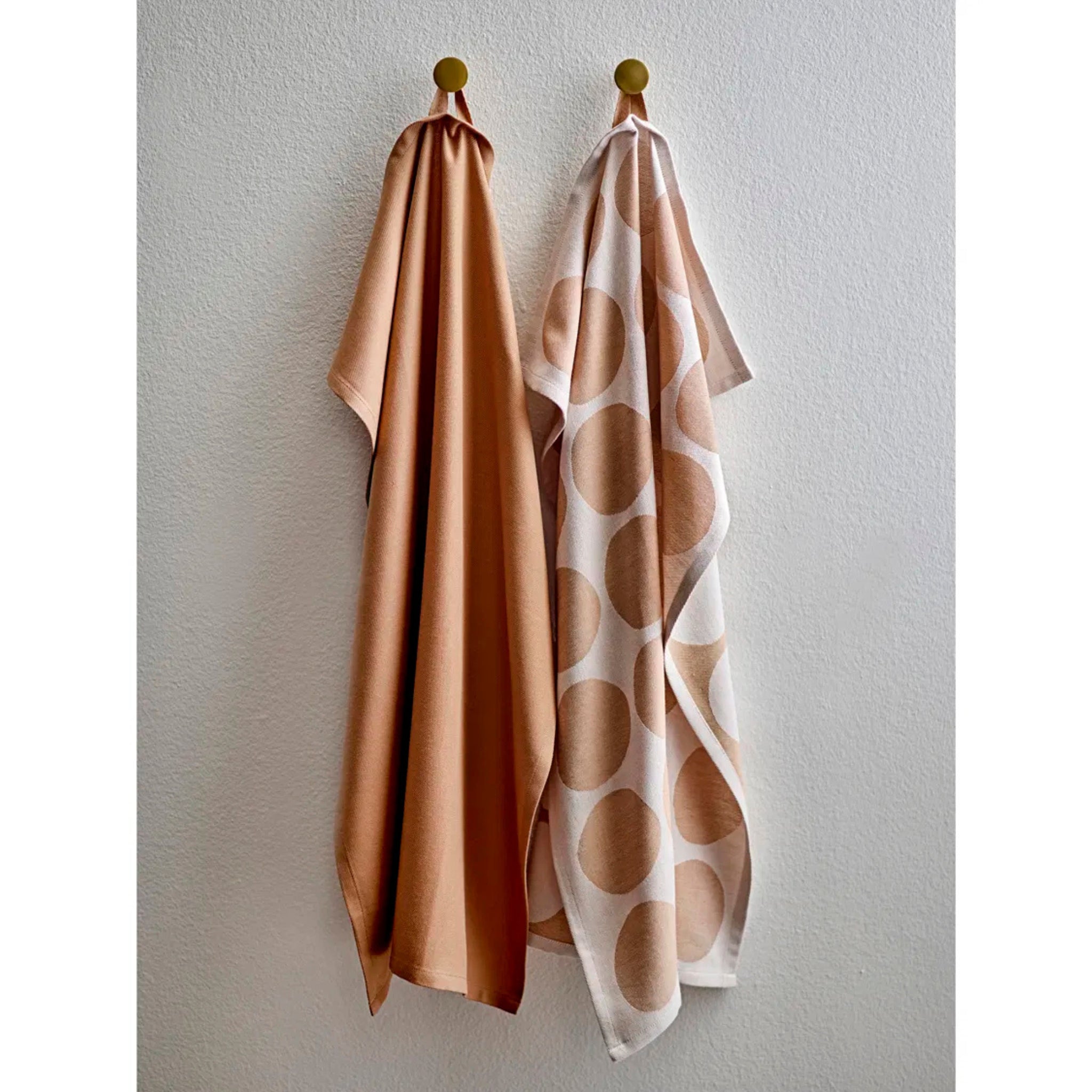Twee Södahl Bubbles theedoeken hangen aan ronde haken tegen een witte muur. De linker is een effen taupe doek, terwijl de rechter wit is met grote tan stippen, beide uit de Sodahl Bubbles Theedoek Taupe 2 st. set.