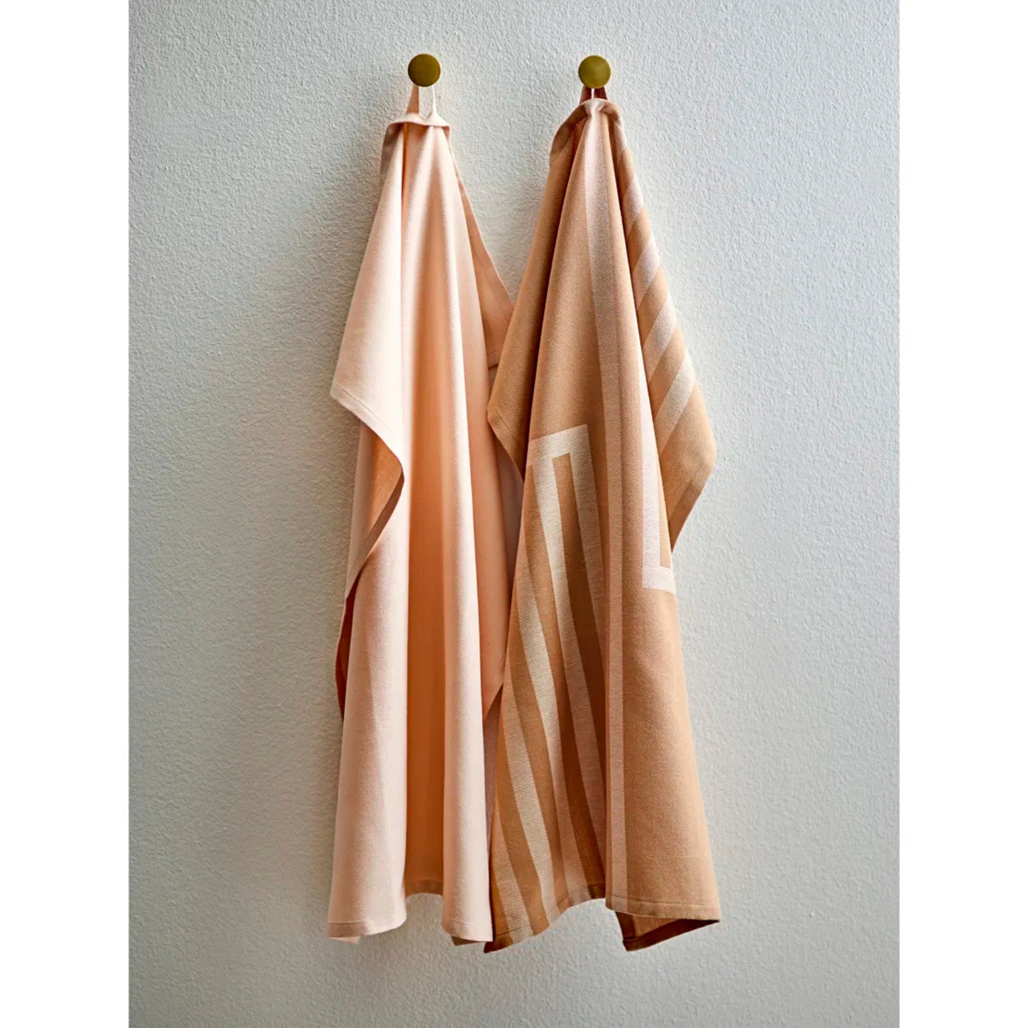 Twee beige theedoeken van Södahl hangen aan gouden haken tegen een lichte muur; de ene is effen taupe en de andere heeft een geometrisch Reflection patroon, beide uit de Sodahl Reflection Theedoek Taupe 2 st. - ideaal voor een Scandinavische keuken.