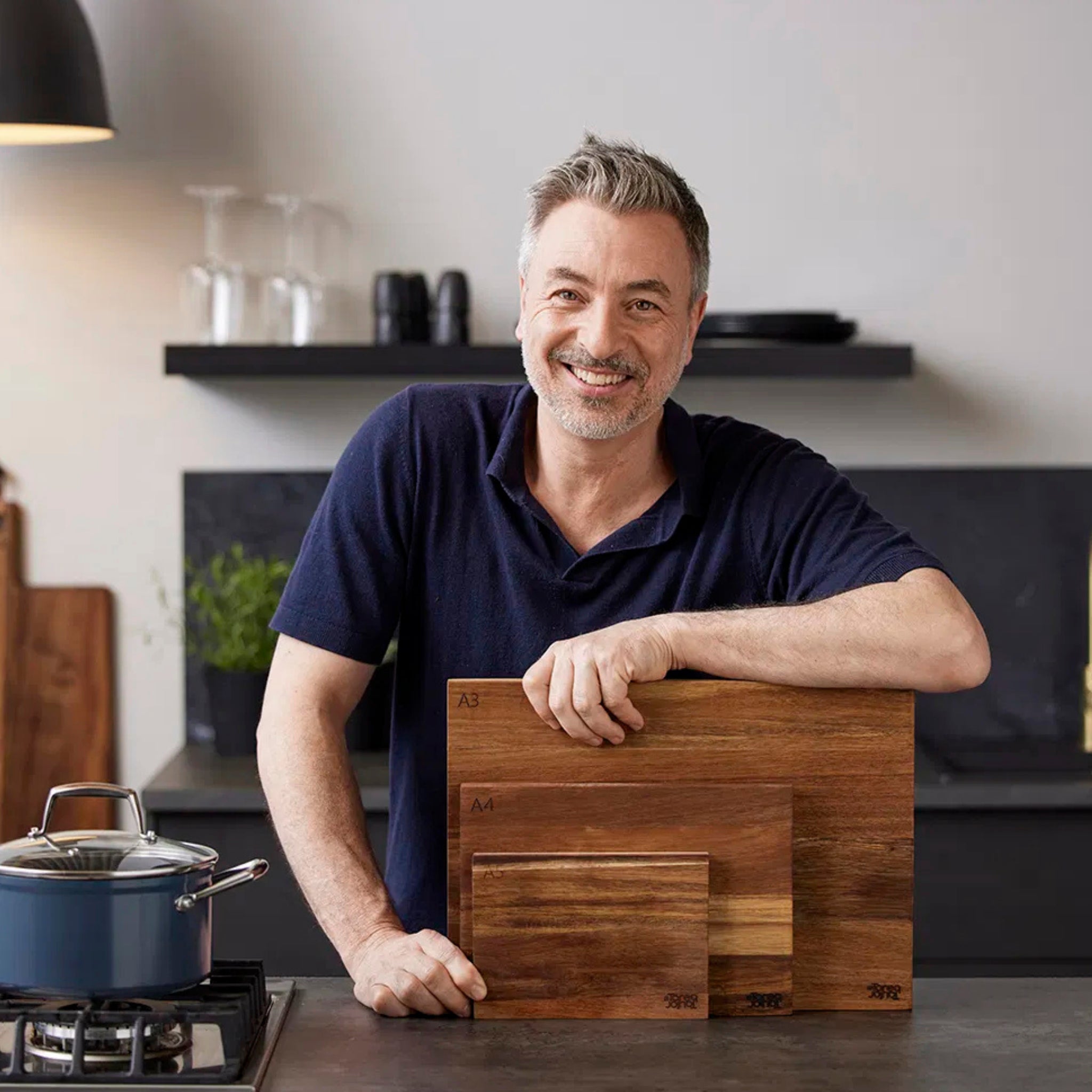 Een lachende man in een marineblauw overhemd staat in een moderne keuken, leunend op het aanrecht terwijl hij drie gestapelde Tareq Taylor Dragon Snijplank Acacia A4 30/21 snijplanken vasthoudt. Vlakbij staat een pan op het fornuis.