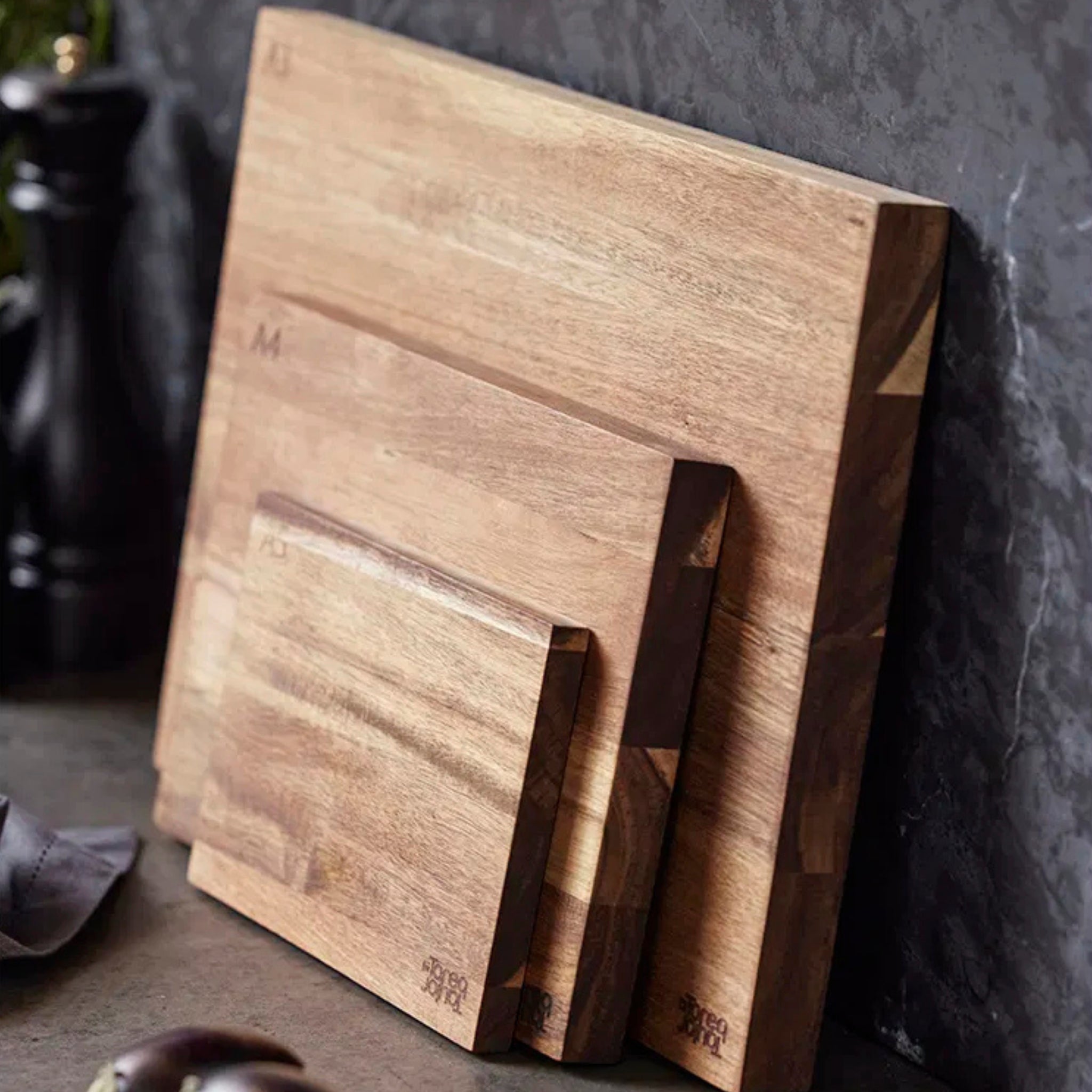 De Tareq Taylor Tarragon Snijplank Acacia A4 30/21 staat rechtop tegen een muur met donkere structuur op een aanrechtblad, met andere keukenartikelen gedeeltelijk zichtbaar op de achtergrond.