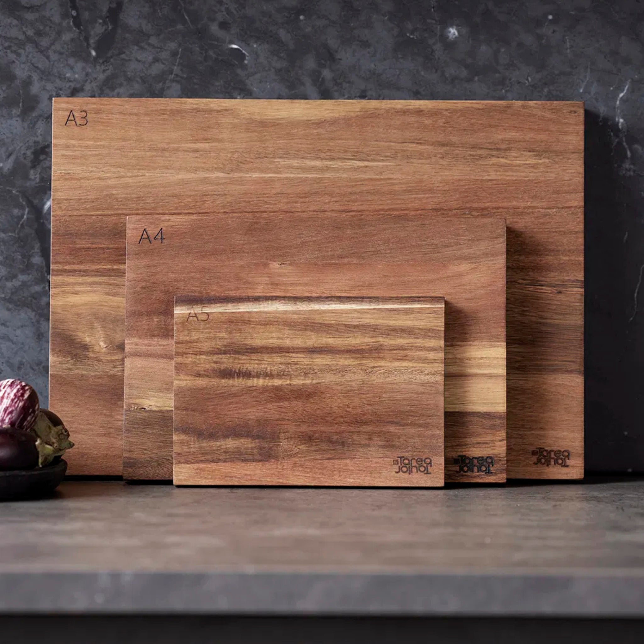 De Tareq Taylor Tarragon Snijplank Acacia A5 21/15 snijplank staat rechtop op een aanrecht tegen een donkere marmeren muur, met een schaal uien in de buurt - een opstelling geïnspireerd door Tareq Taylor.