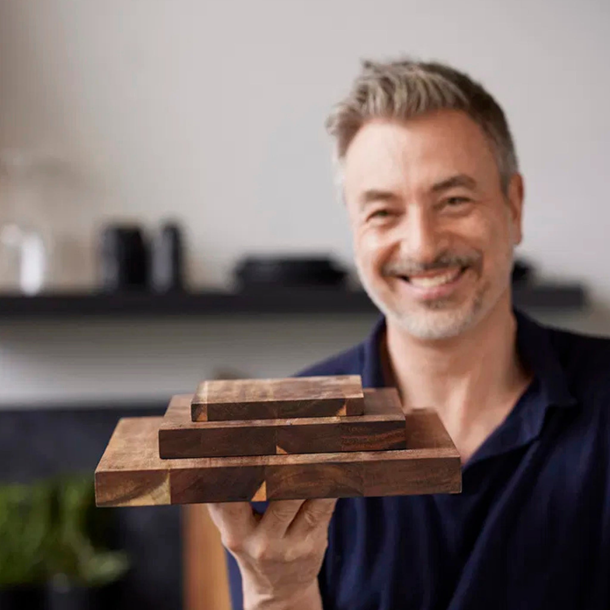 Een lachende man met grijs haar en een baard houdt een stapel houten blokken omhoog, waaronder de Tareq Taylor Tarragon Snijplank Acacia A4 30/21 van Tareq Taylor. De zacht vervaagde achtergrond toont een gezellige kamer met planken en planten.