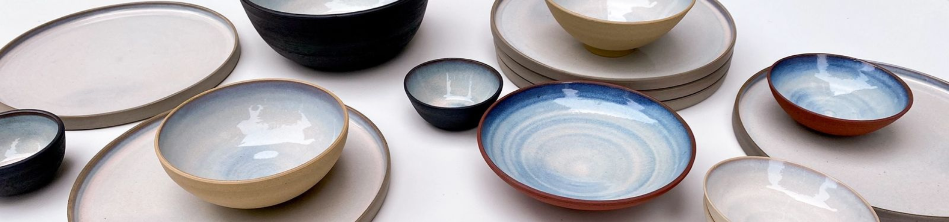 Verschillende handgemaakte keramische kommen en borden met neutrale en blauwe tinten zijn gerangschikt op een wit oppervlak, waarbij verschillende maten en vormen met een minimalistische, moderne esthetiek worden getoond.
