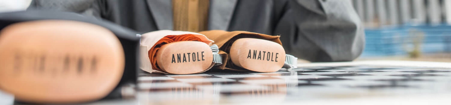 Close-up van drie gevouwen Anatole paraplu's op een geruit oppervlak, met de houten handvaten naar voren en een persoon vaag op de achtergrond.