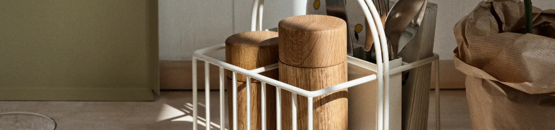 In een witte metalen caddy staan twee houten bakjes, keukengerei en keukenspullen op een licht oppervlak. Een bruine papieren zak ligt vlakbij in een zonnige keukenomgeving.