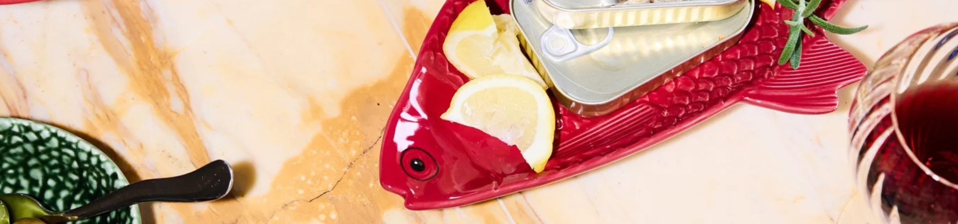 Op een rood visvormig bord liggen citroenschijfjes en een open blikje sardines. Het bord staat op een marmeren oppervlak naast een glas rode drank en een deel van een groene schaal met een lepel.