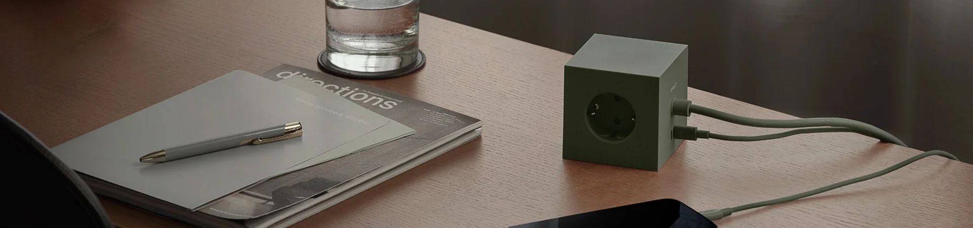Een groene kubusvormige stekkerdoos met twee snoeren erin ligt op een houten bureau naast een notitieblok, pen, tijdschrift, glas water en een smartphone.