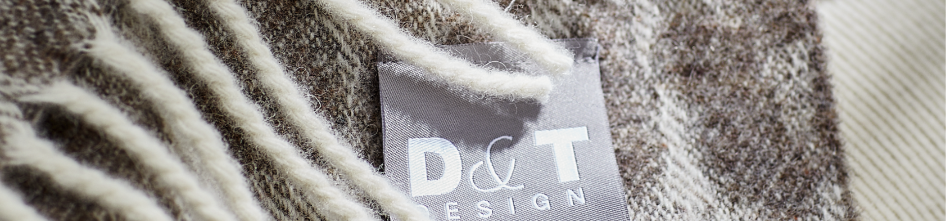Close-up van een grijs stoffen label met D&T DESIGN tekst, bevestigd aan een wollen kledingstuk met zachte, crèmekleurige franjes en een bruin en beige structuurpatroon.