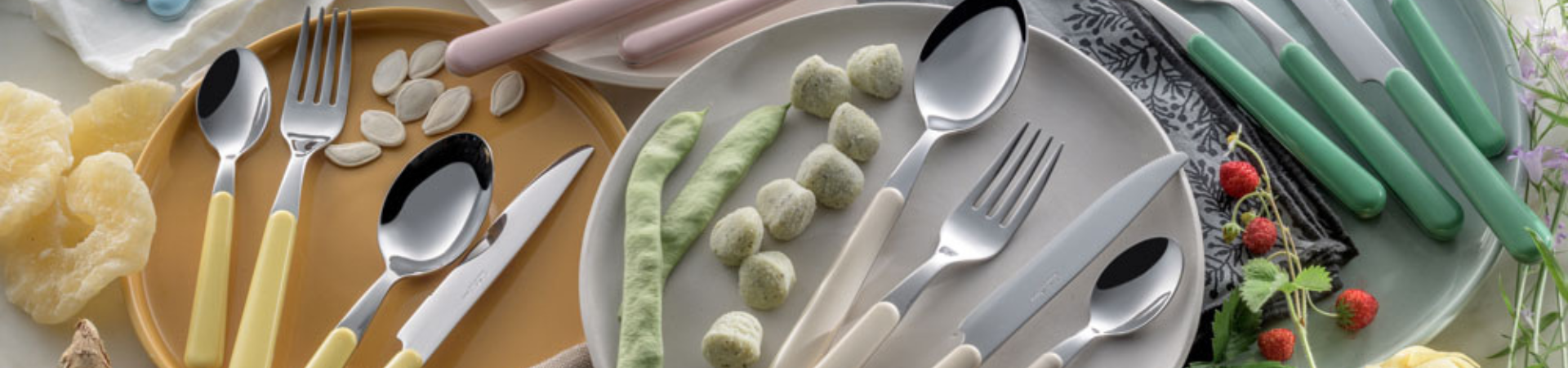 Assortiment bestek met pastelkleurig handvat, waaronder lepels, vorken en messen, gerangschikt op borden met etenswaren zoals gnocchi, kaas en zaden, omringd door bloemen en bessen.