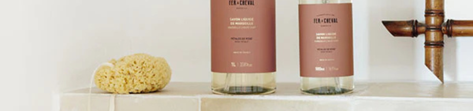 Twee flessen vloeibare zeep met roze labels en een natuurlijke zeespons staan op een betegelde badkamerplank naast een bronzen kraan.