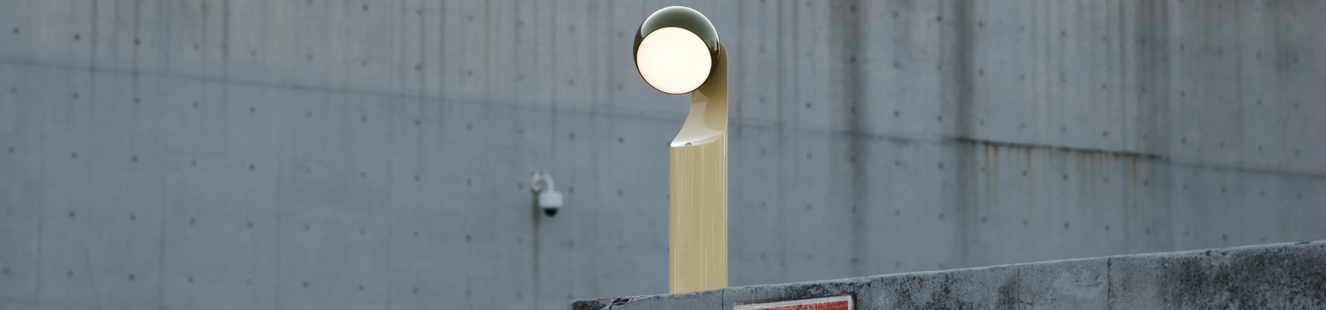 Een moderne straatlantaarn met een ronde lamp staat vlakbij een betonnen muur. Op de achtergrond is een beveiligingscamera te zien en het oppervlak is bezaaid met kleine gaatjes.