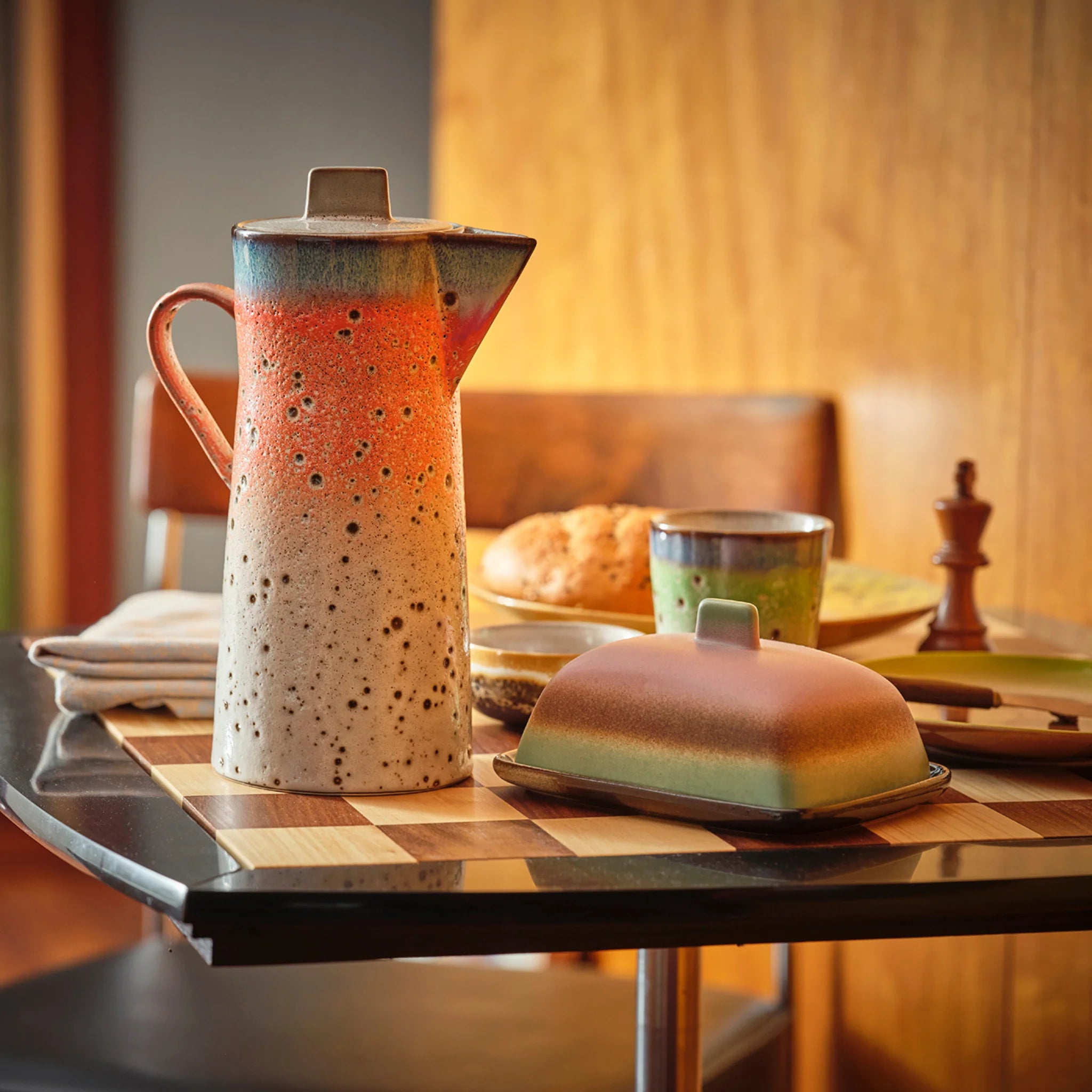 Een keramische kruik en botervloot met een gespikkeld, gradiëntglazuur staan op een ontbijttafel gedekt met brood, een kopje, servetten en een pepermolen in een gezellige, warm verlichte kamer.