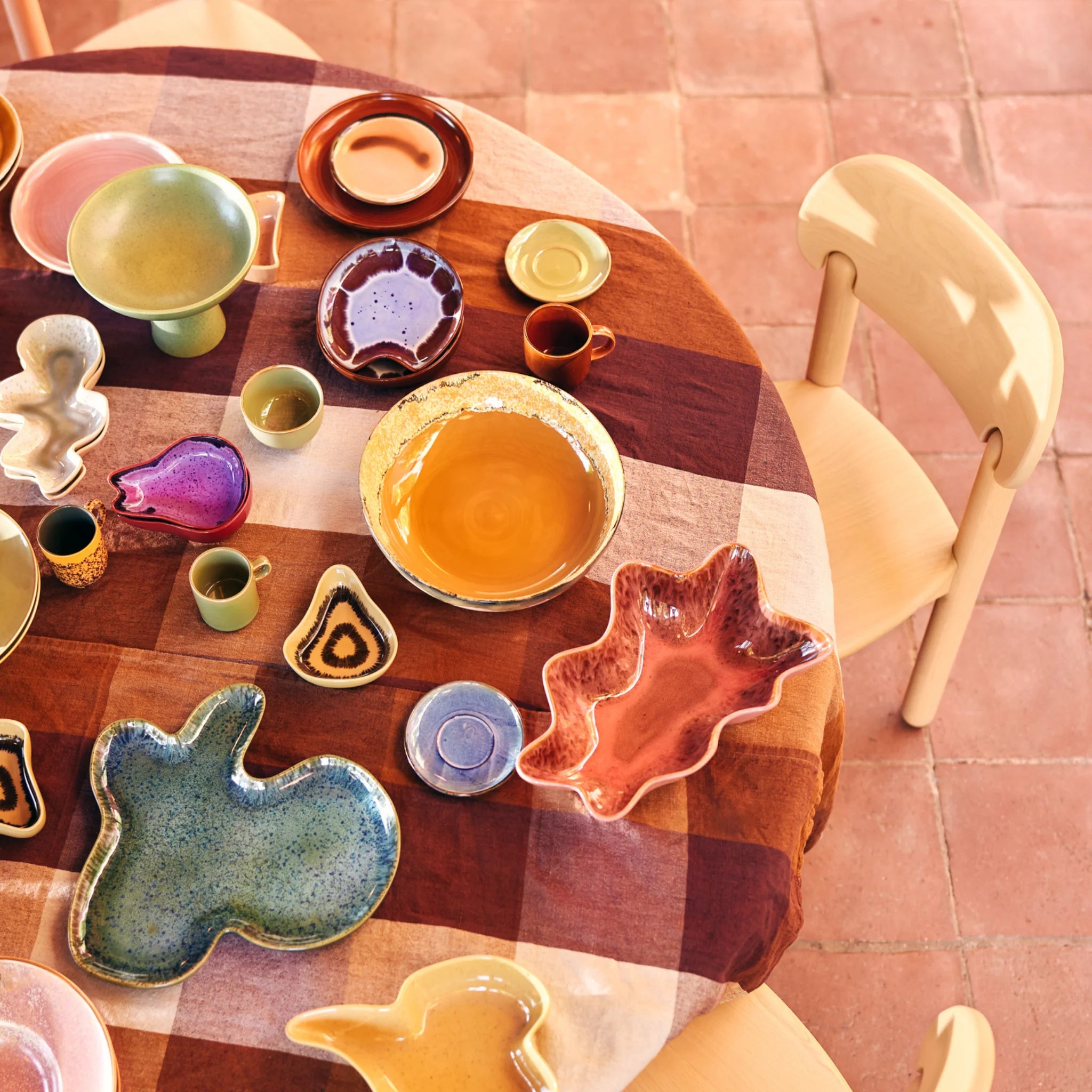 Een gedekte tafel met kleurrijke, uniek gevormde keramische borden en kopjes staat op een rode tegelvloer, naast een houten stoel. Het geruite tafelkleed heeft bruine en beige tinten.