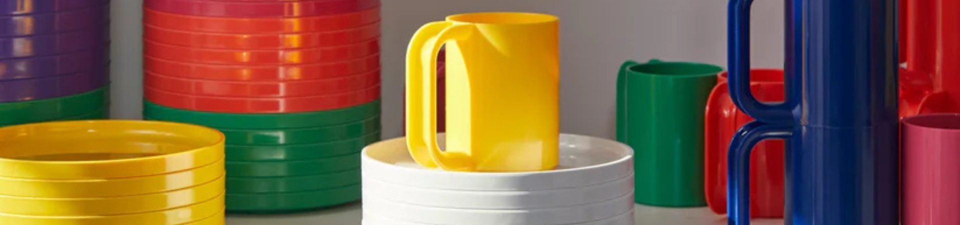 Kleurrijk plastic servies, waaronder gestapelde borden, kommen en mokken in geel, rood, groen, roze en blauw, netjes gerangschikt op een plank.