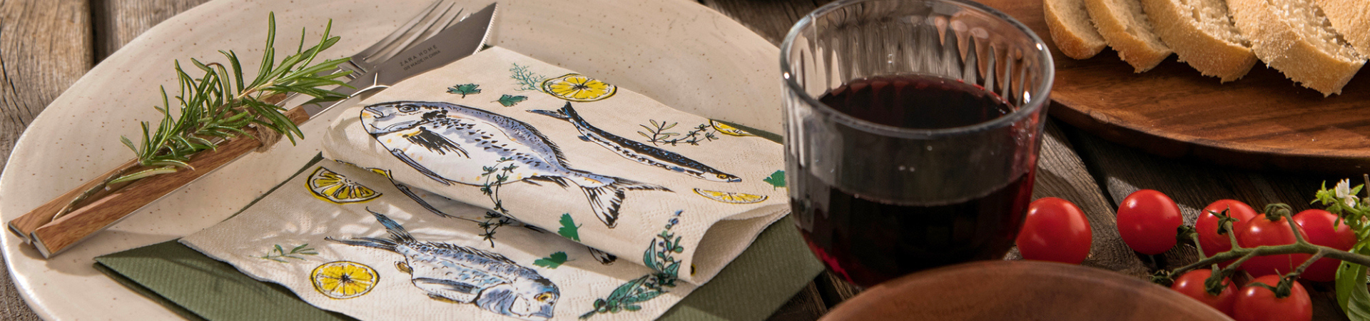 Een gedekte tafel met een bord, een vork met rozemarijn, een servet met vismotief, een glas rode wijn, gesneden brood en kerstomaatjes op een rustiek houten oppervlak.