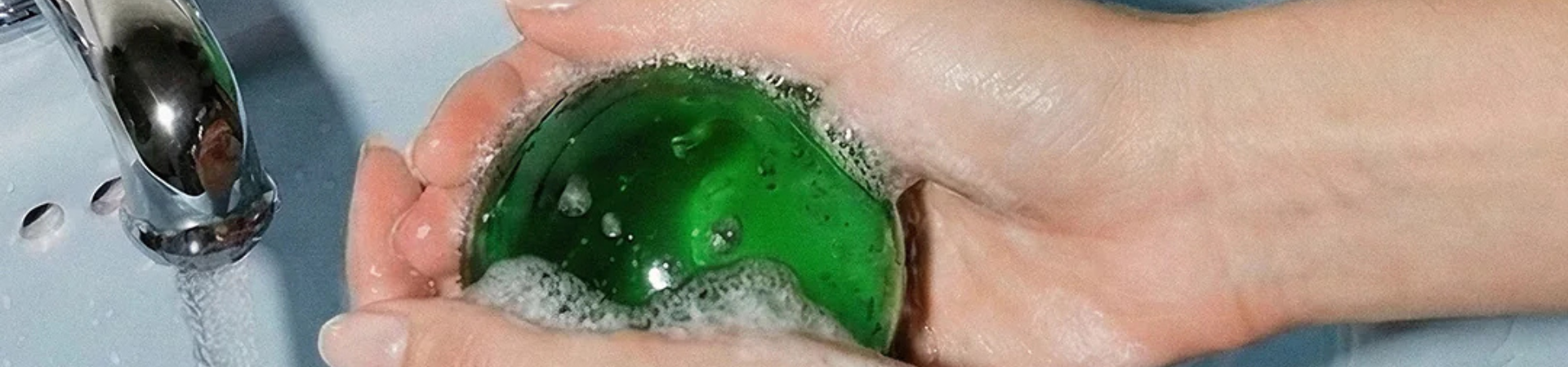 Handen die een ronde groene zeep onder stromend water in een wasbak wassen, waarbij er belletjes op de zeep ontstaan.