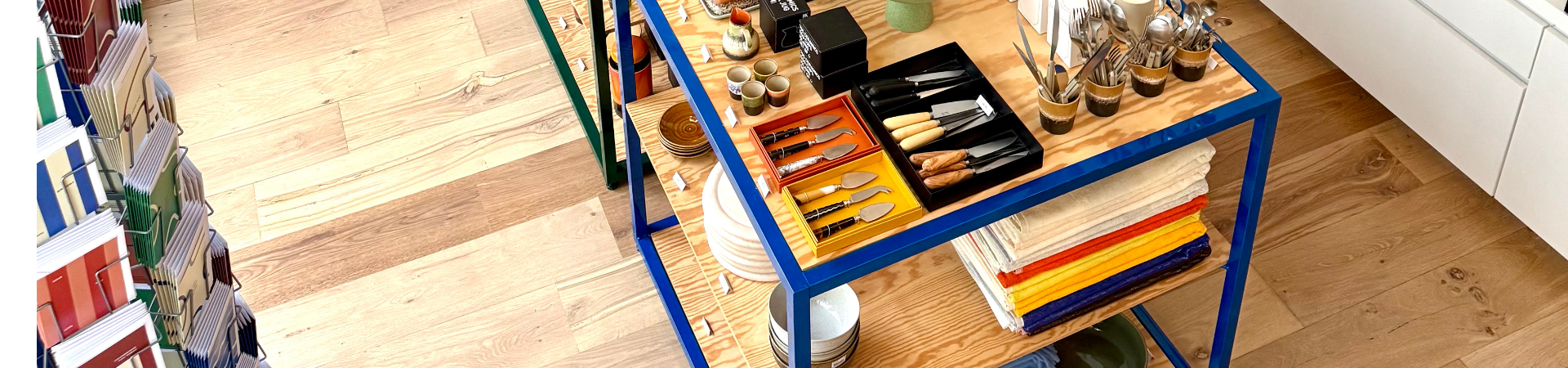 Een winkeldisplay met keukengerei, kleurrijke servetten, messen, kopjes en borden netjes gerangschikt op houten planken met een blauw metalen frame, op een lichte houten vloer.