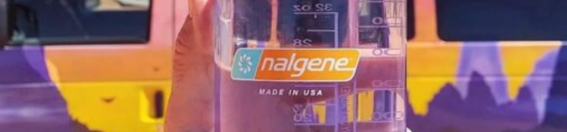 Een hand houdt een heldere Nalgene waterfles gevuld met water vast; een kleurrijk busje met paarse en oranje bergmotieven staat op de wazige achtergrond.