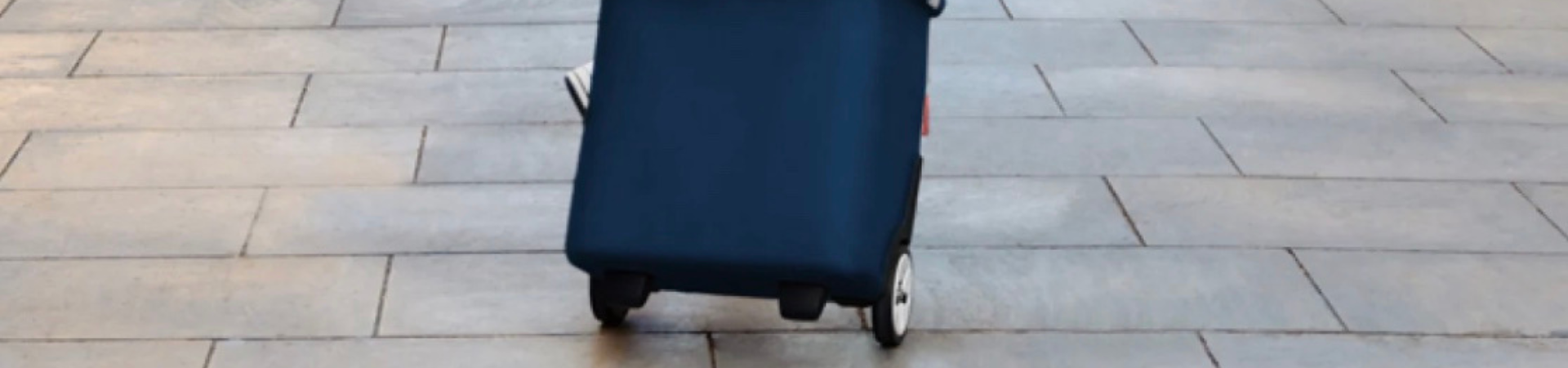 Een blauwe koffer op wieltjes staat rechtop op een lichtgrijs betegeld oppervlak, van voren gezien zonder zichtbare handvatten of achtergrondelementen.