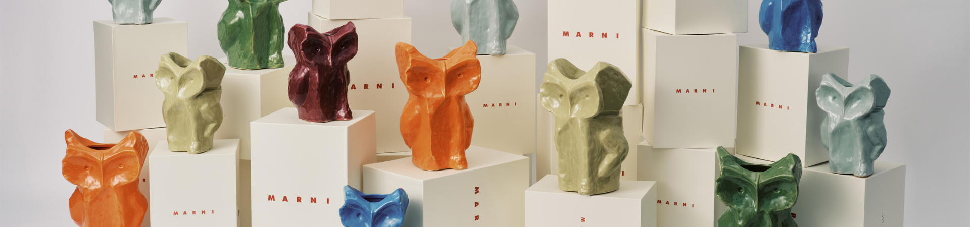 Kleurrijke keramische sculpturen in de vorm van dieren, in de kleuren oranje, groen, blauw en lichtgrijs, worden tentoongesteld op witte dozen van het merk Marni, die op verschillende hoogtes tegen een effen achtergrond zijn opgesteld.