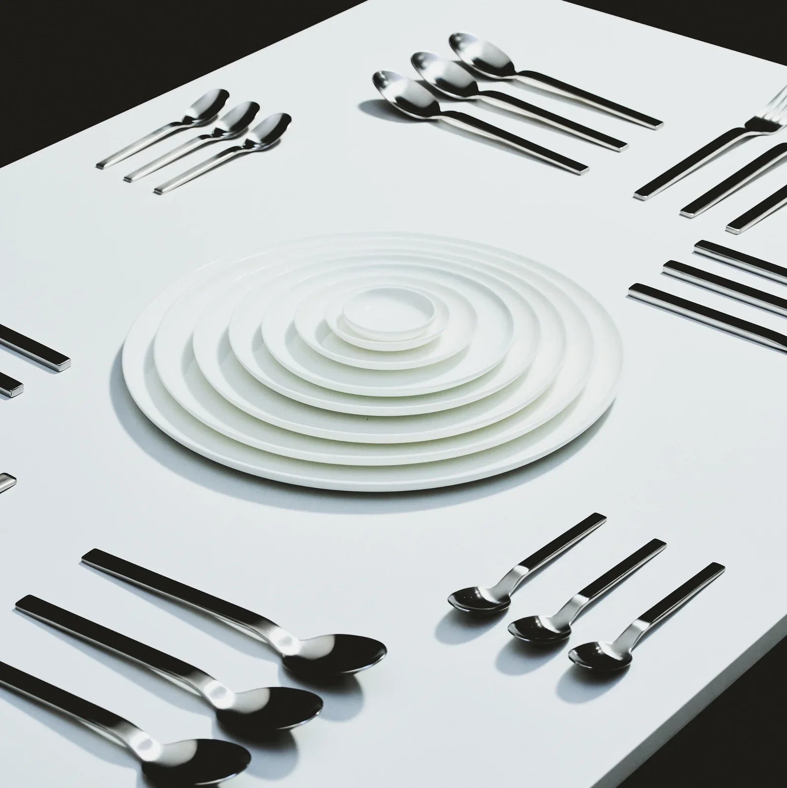 Een witte tafel toont keurig gerangschikte sets zilverwerk en gestapelde witte borden van verschillende grootte, waardoor een symmetrisch en georganiseerd patroon ontstaat.