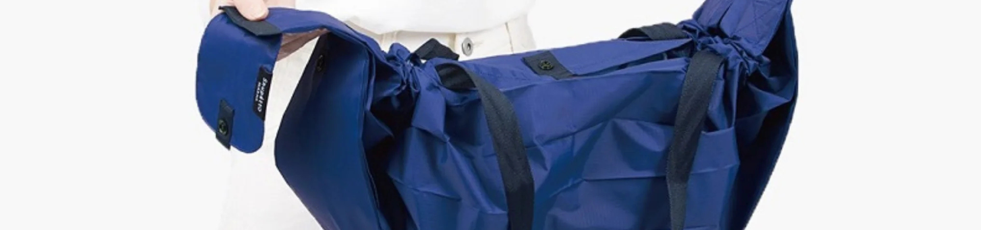 Een persoon in een witte outfit houdt een grote blauwe draagtas met zwarte riemen en trekkoorden aan de zijkant open, waardoor de ruime binnenkant zichtbaar wordt.