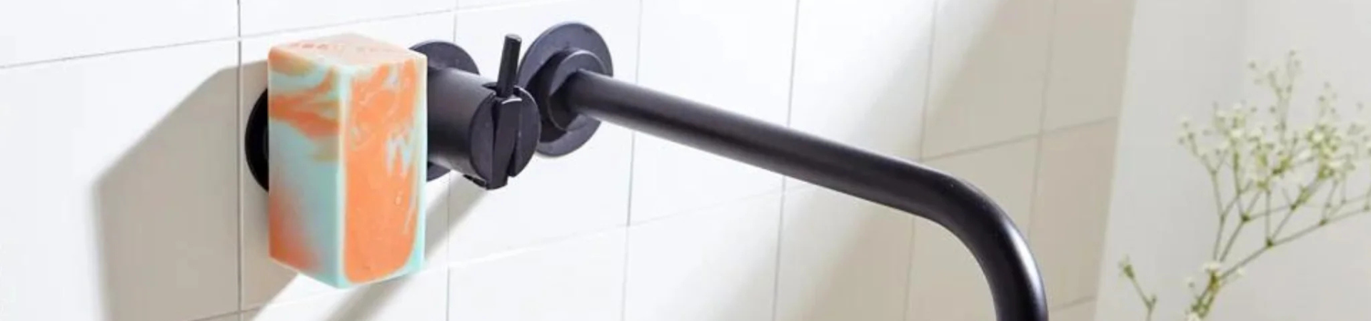 Een gestreept stuk zeep is gemonteerd op een magnetische houder die is bevestigd aan een wit betegelde muur boven een zwart metalen handdoekenrek, met enkele groene plantenstengels zichtbaar op de achtergrond.