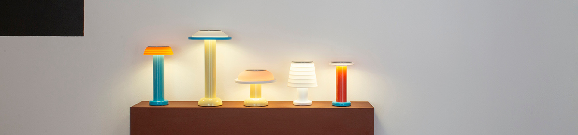 Vijf kleurrijke moderne tafellampen van verschillende hoogtes en ontwerpen staan op een rij op een bruine console tegen een effen witte muur en werpen zacht licht.