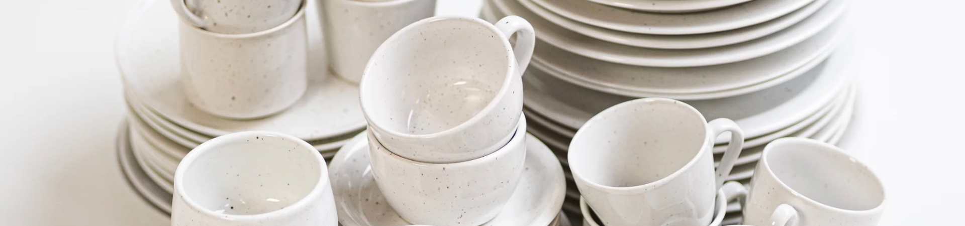 Een verzameling gestapelde witte keramische kopjes, schoteltjes en borden met een gespikkeld patroon, netjes gerangschikt tegen een witte achtergrond.