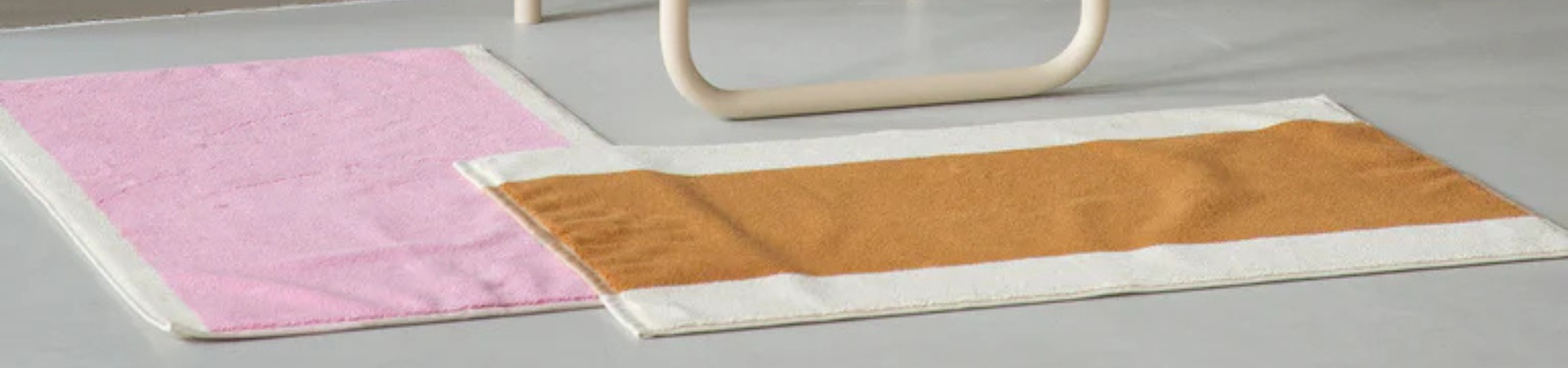 Op een grijze vloer liggen twee rechthoekige vloerkleden: het ene is effen roze en het andere heeft een brede taankleurige streep omzoomd door wit. Op de achtergrond is een gedeelte van gebogen, lichtgekleurd meubilair zichtbaar.
