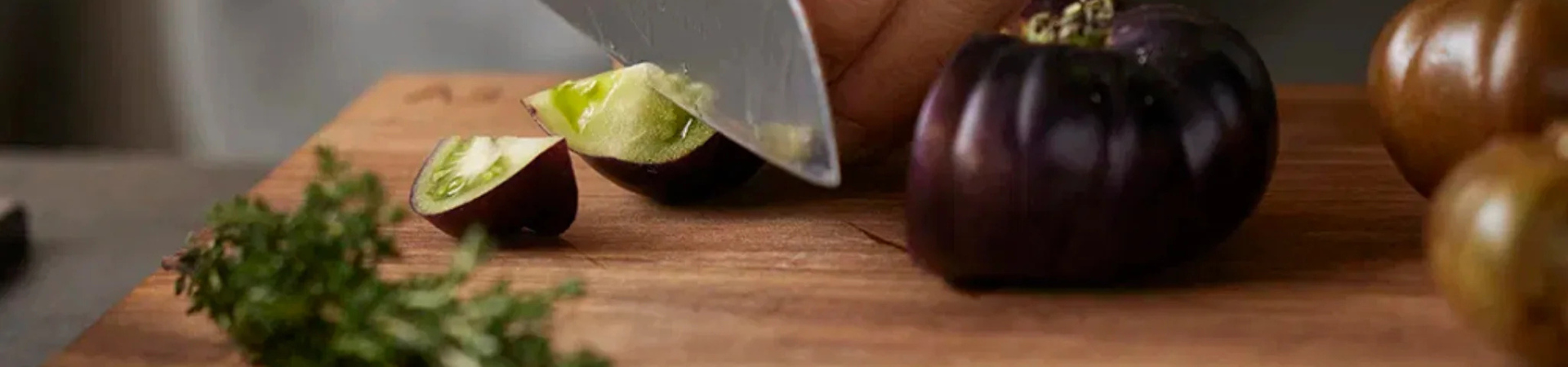Een close-up van een hand die een aubergine snijdt op een houten snijplank, met verse kruiden en andere groenten in de buurt.