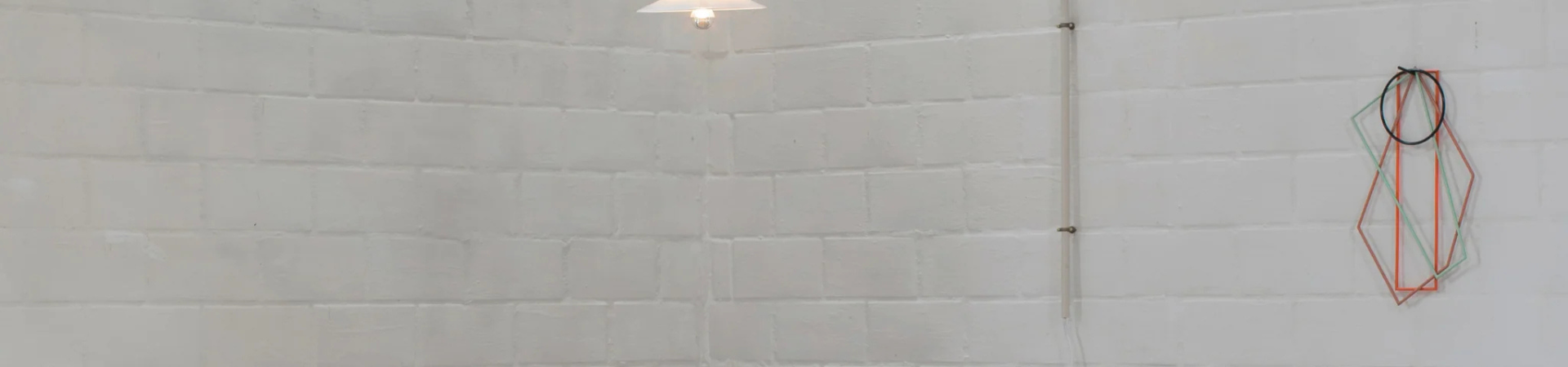 Minimalistische witte bakstenen muur met een hanglamp in de hoek en geometrische draadkunstwerken in zwart, rood en groen aan de rechterkant. De kamer heeft een strakke, moderne uitstraling.